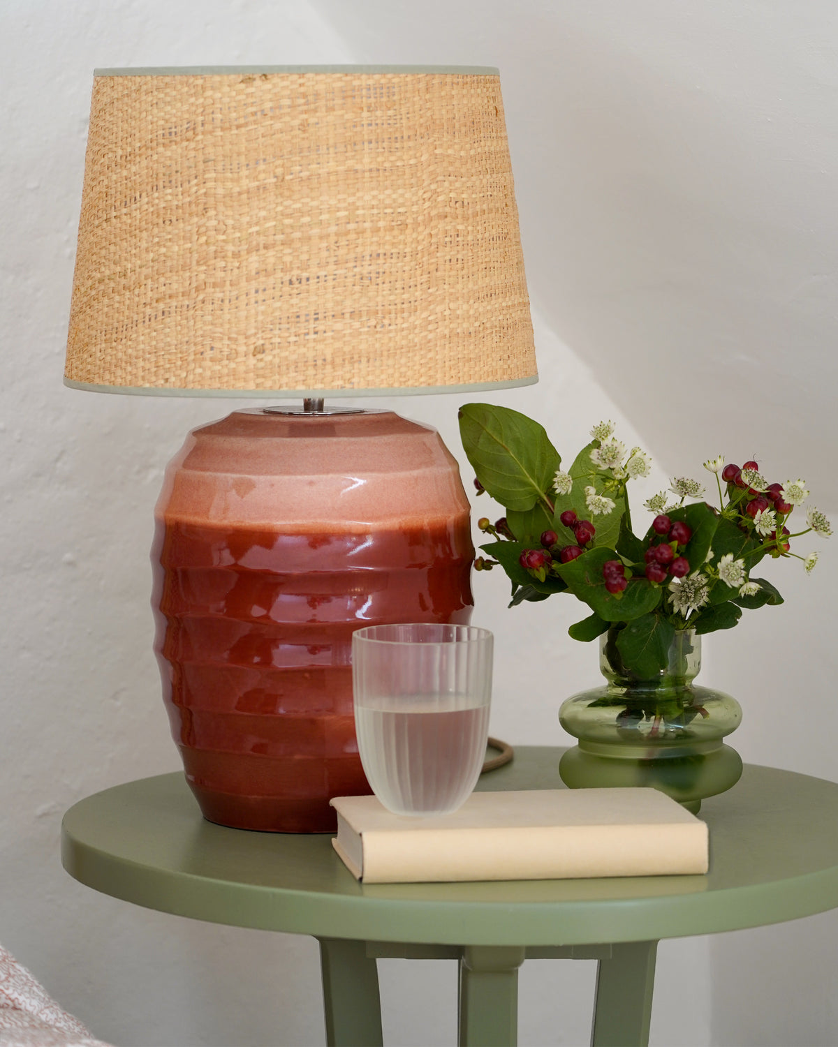 Decorative green table with a red ceramic lamp, glass, book, and small plant on a light background