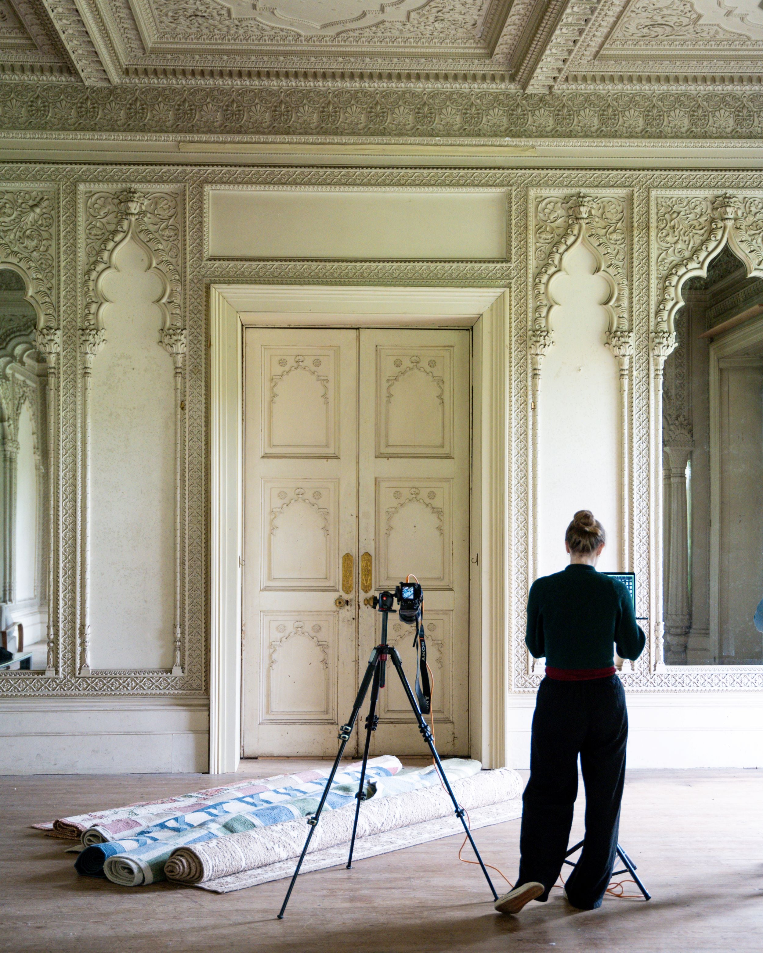 Person setting up camera equipment in an ornate room with decorative walls and a large door.