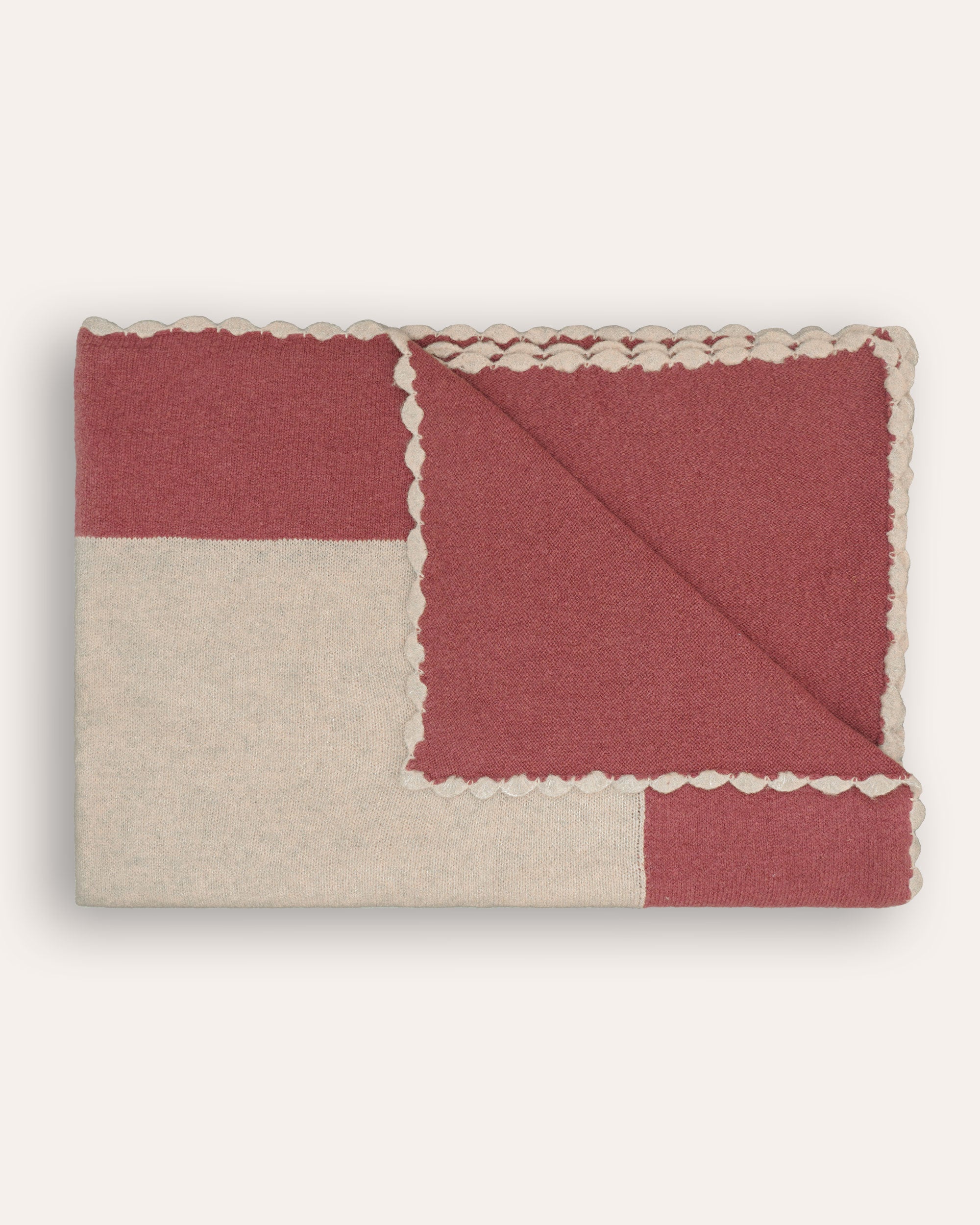 A red and beige knitted throw with a scalloped edge design, laid out on a plain background.