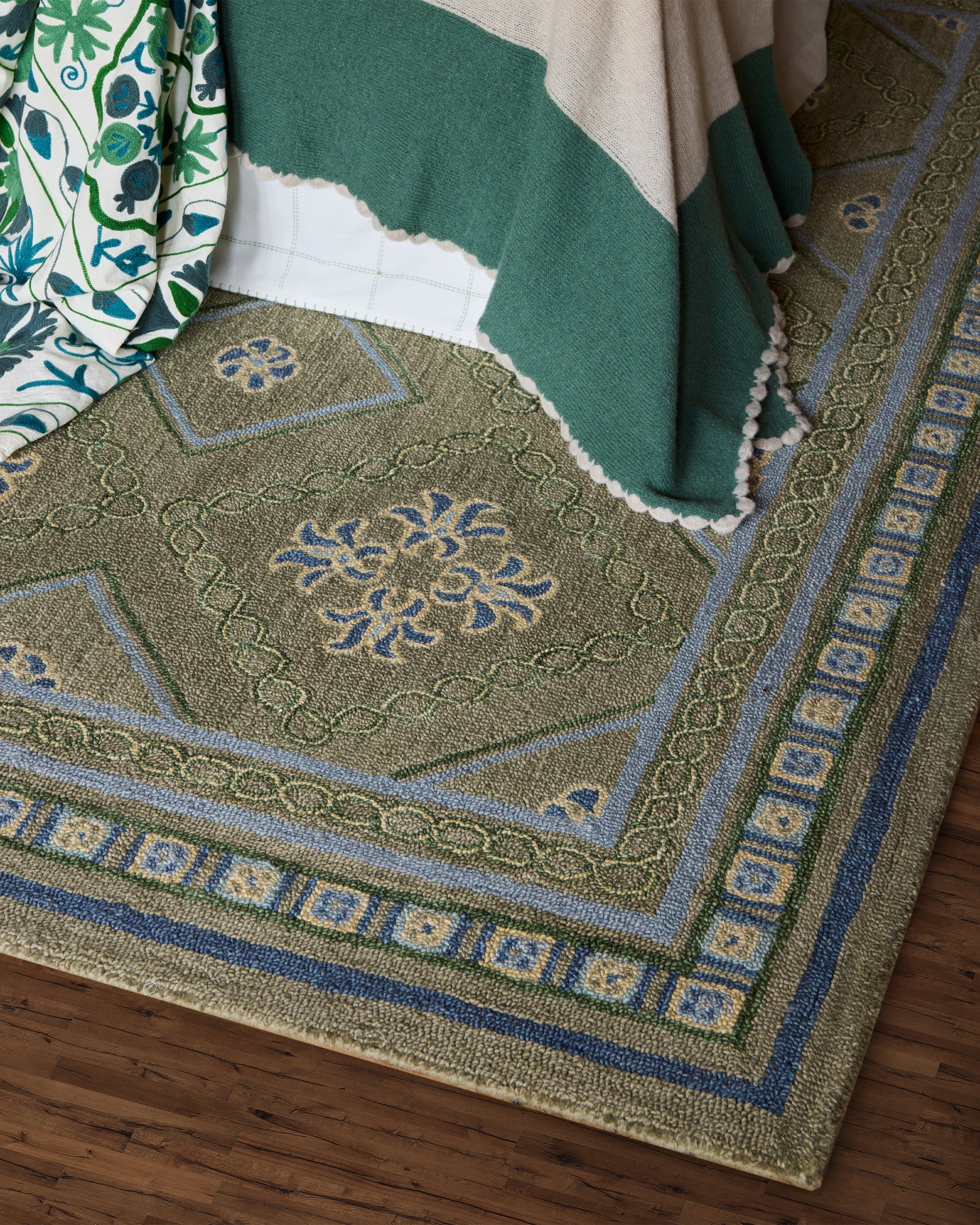 Green patterned Varuna rug on a wooden floor with a blanket draped over it.