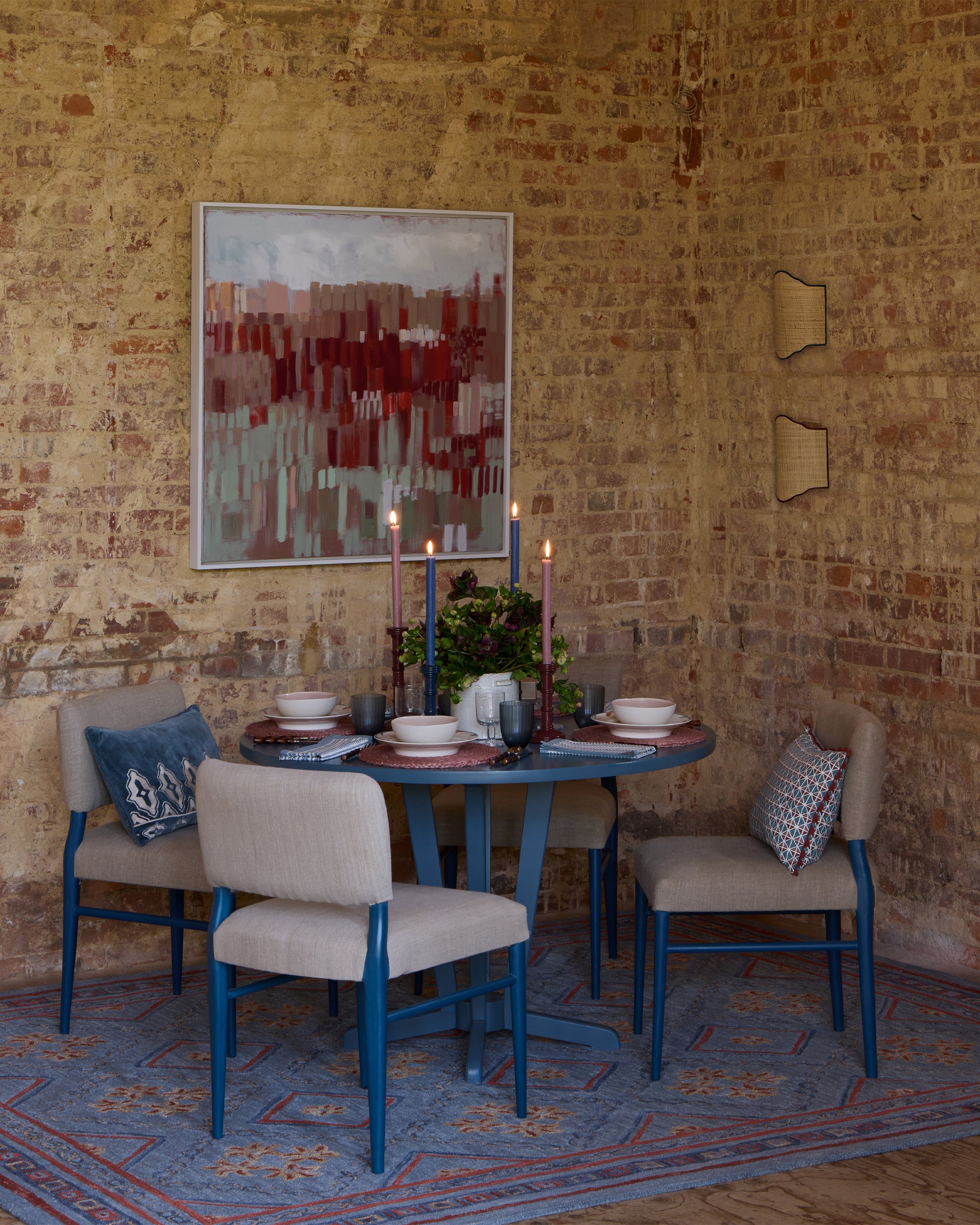 Dining area with a round table and chairs against a brick wall with a painting.