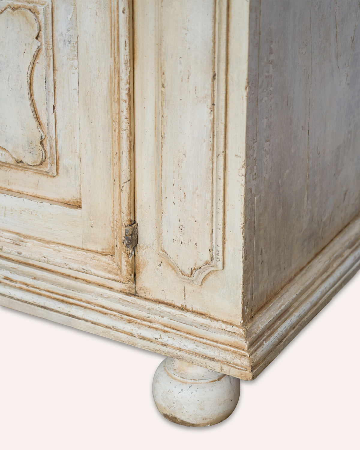 Close-up of a distressed vintage-style cabinet corner, showing carved panel details, a turned wooden foot, and a weathered off-white finish with subtle warm beige undertones.