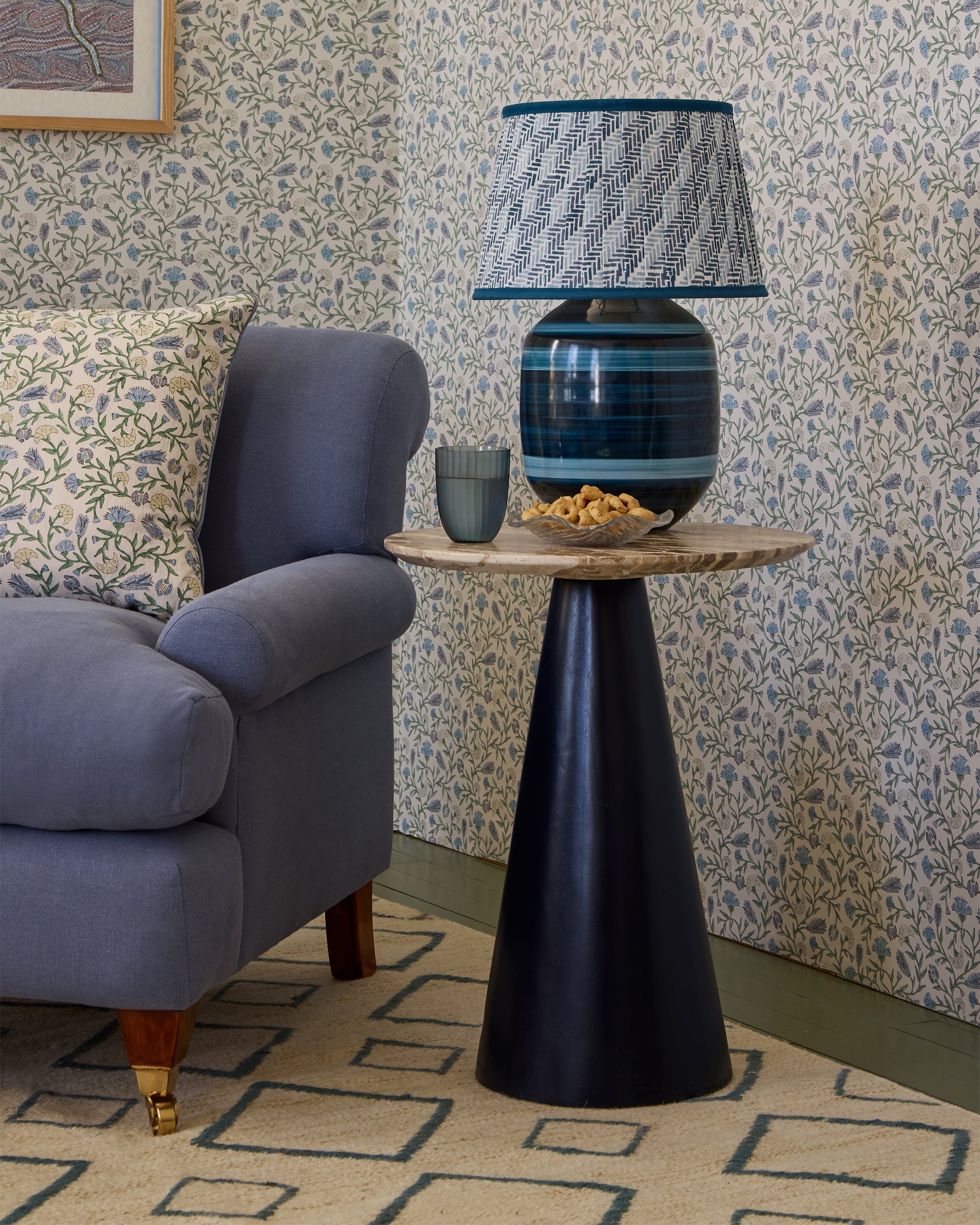 Decorative lamp with striped design on a side table in a room with floral wallpaper and a blue armchair.