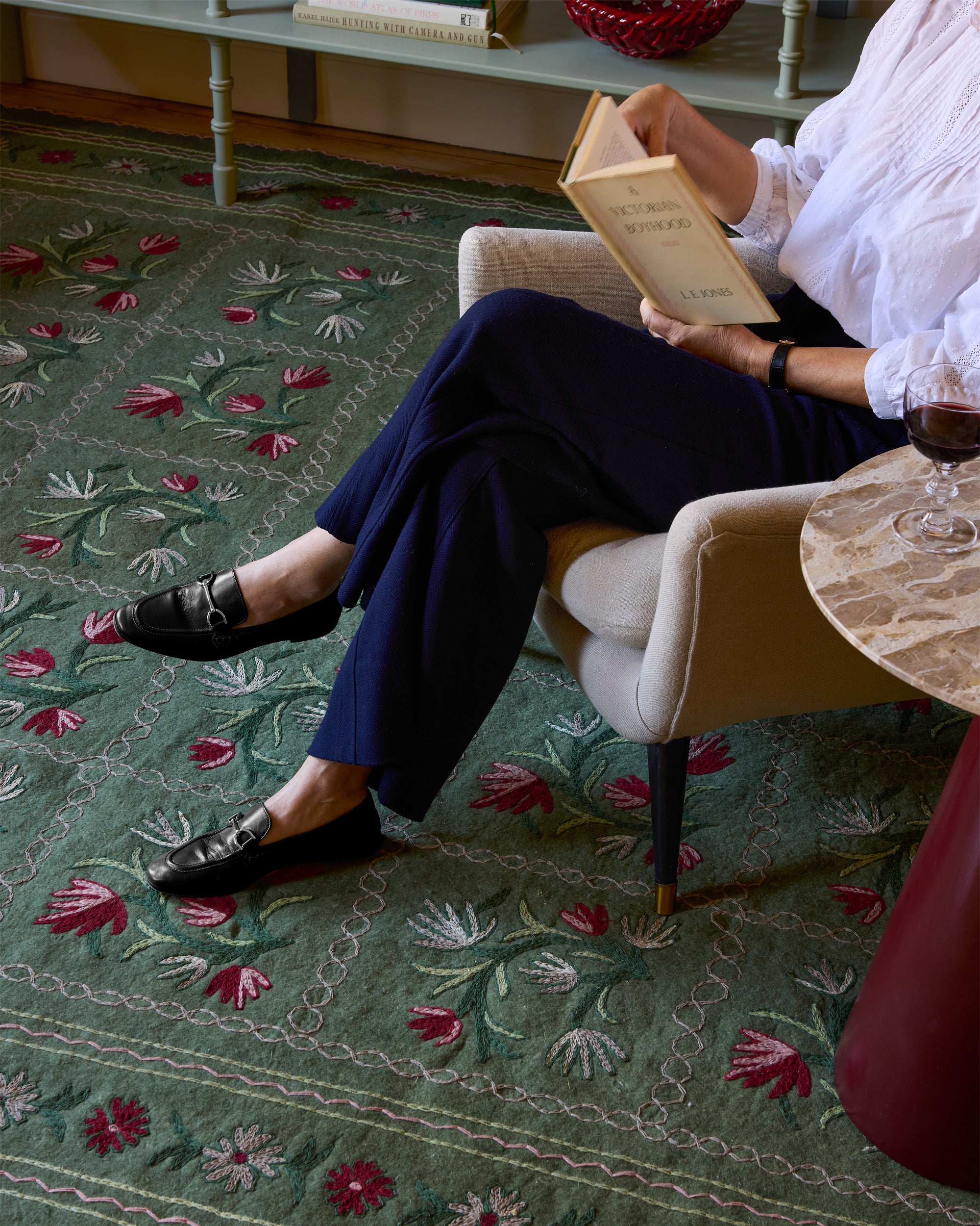 Person sitting on a chair reading a book on a patterned rug with a table and wine glass in the background.