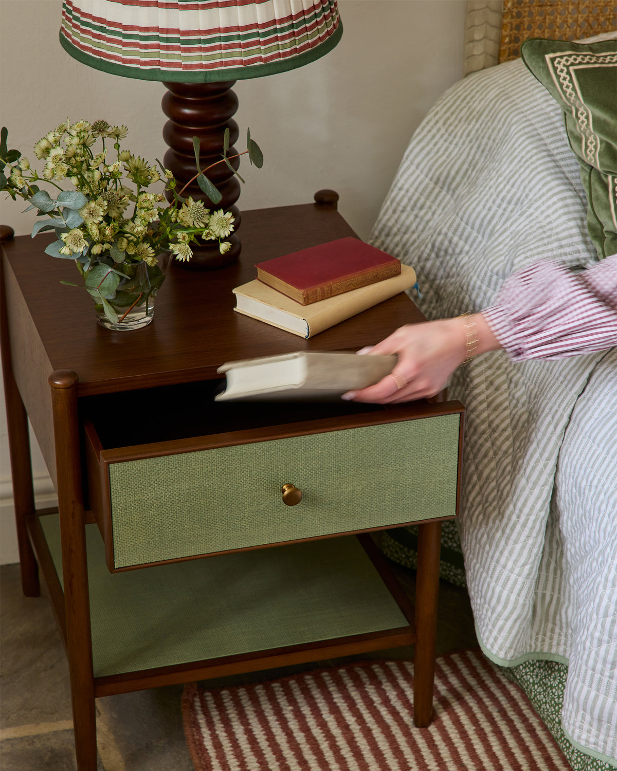 Person holding a book next to a wooden side table with a lamp and books on it.