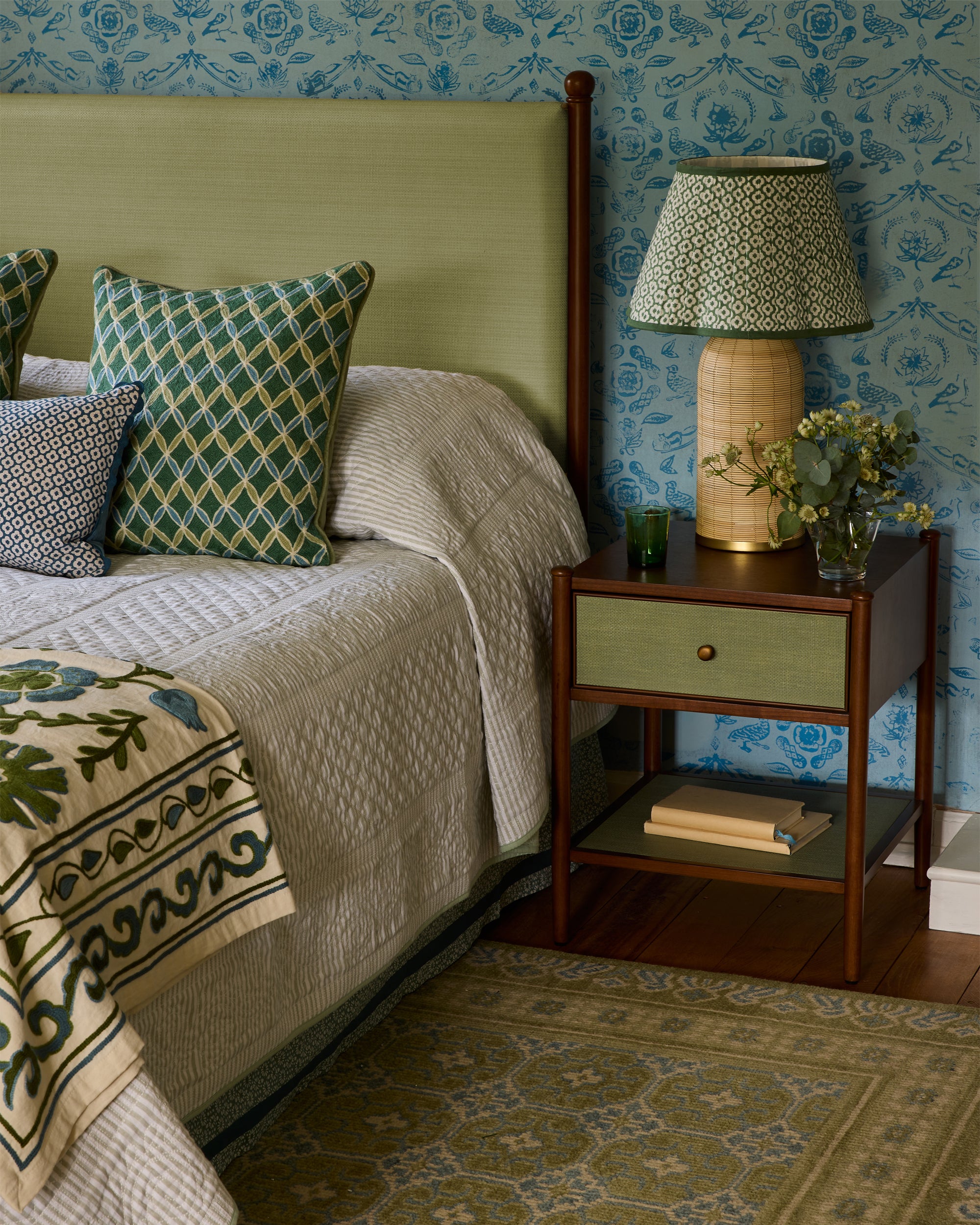 Bedroom with green and beige bedding, patterned wallpaper, and a nightstand with a lamp.