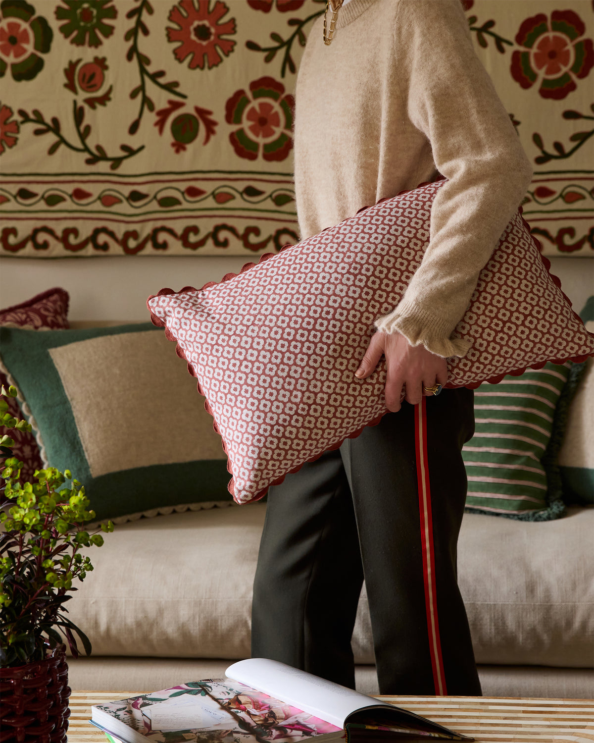 Person holding a patterned cushion in a sitting room with decorative cushions and a colourful wall.