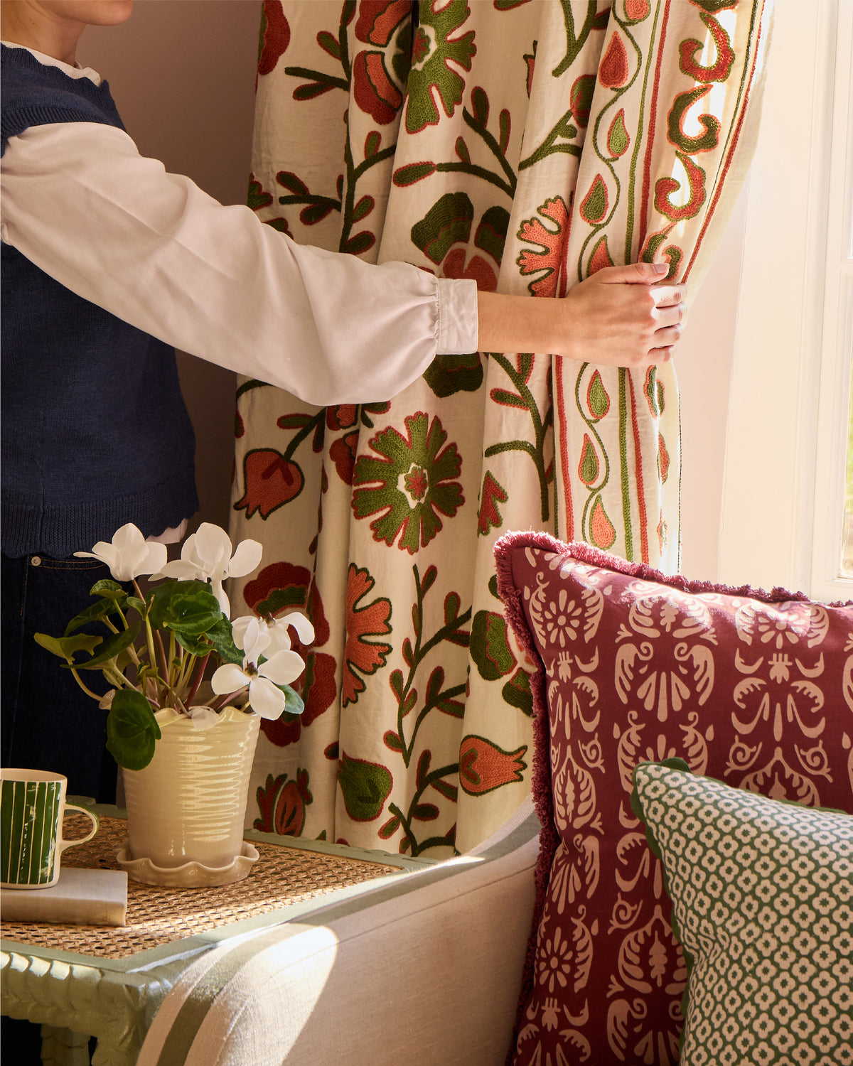 Person adjusting a patterned curtain in a room with decorative cushions and a plant.