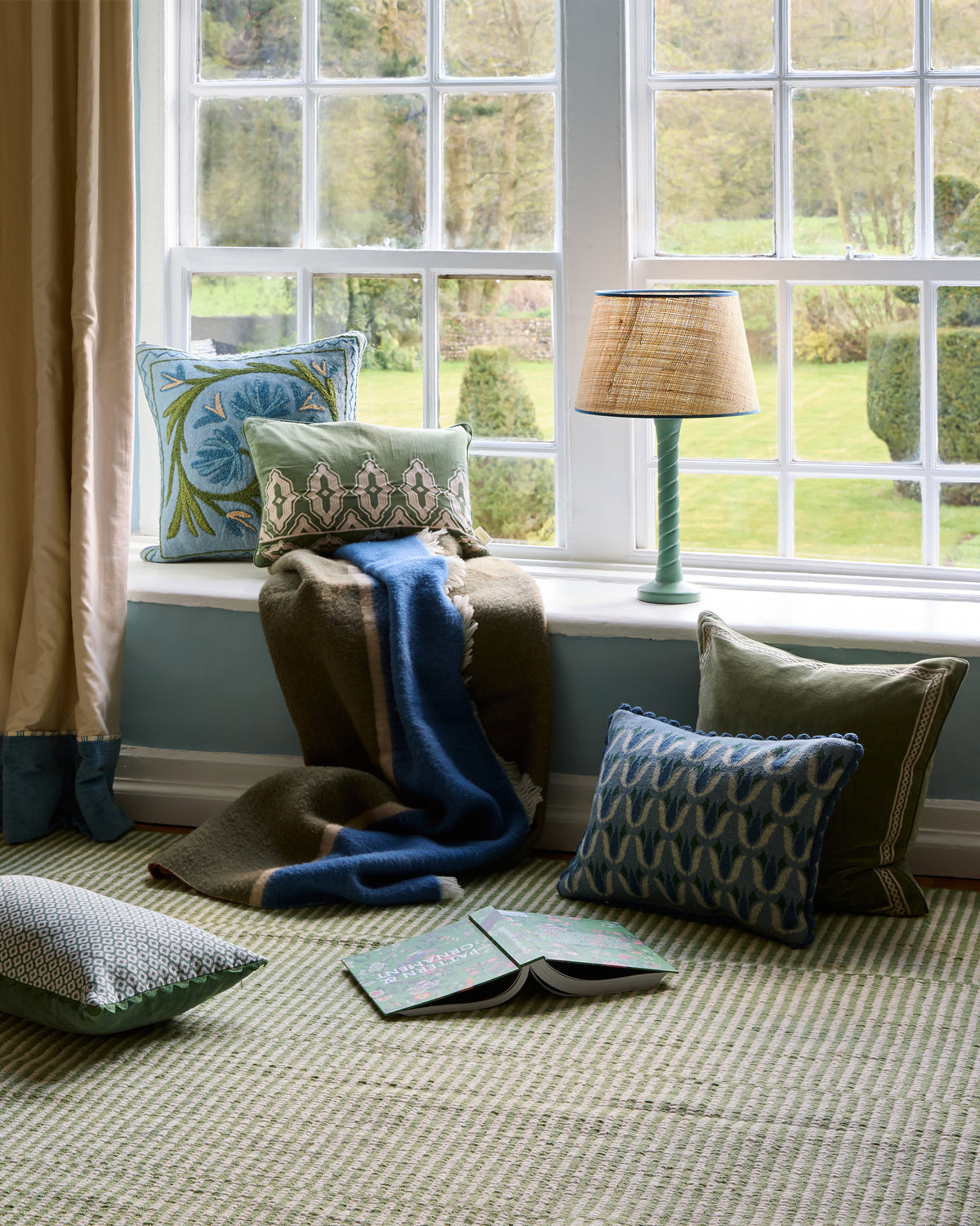 Cozy reading nook with cushions, a blanket, and a lamp by a window with a view of greenery.