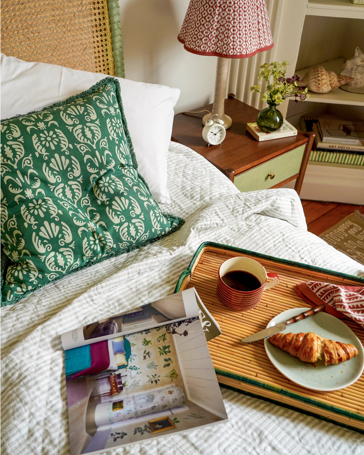 A styled bedroom scene with a green damask-patterned throw pillow on a bed with white bedding. A bamboo tray on the bed holds a cup of coffee, a croissant on a plate, and a magazine.