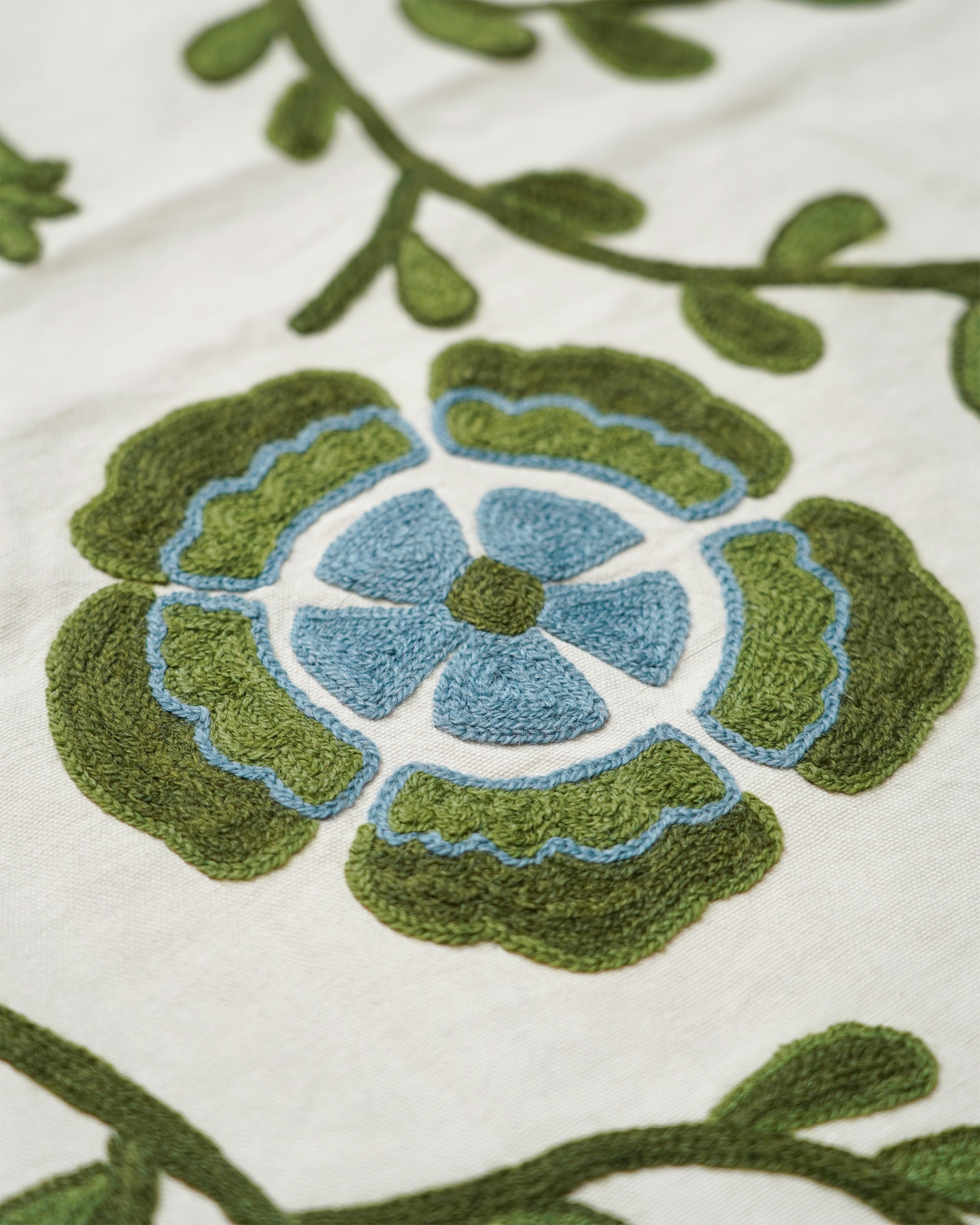 Close-up of a floral embroidered textile design featuring a blue flower center surrounded by layered green petals and olive-green vine outlines on a white fabric background.