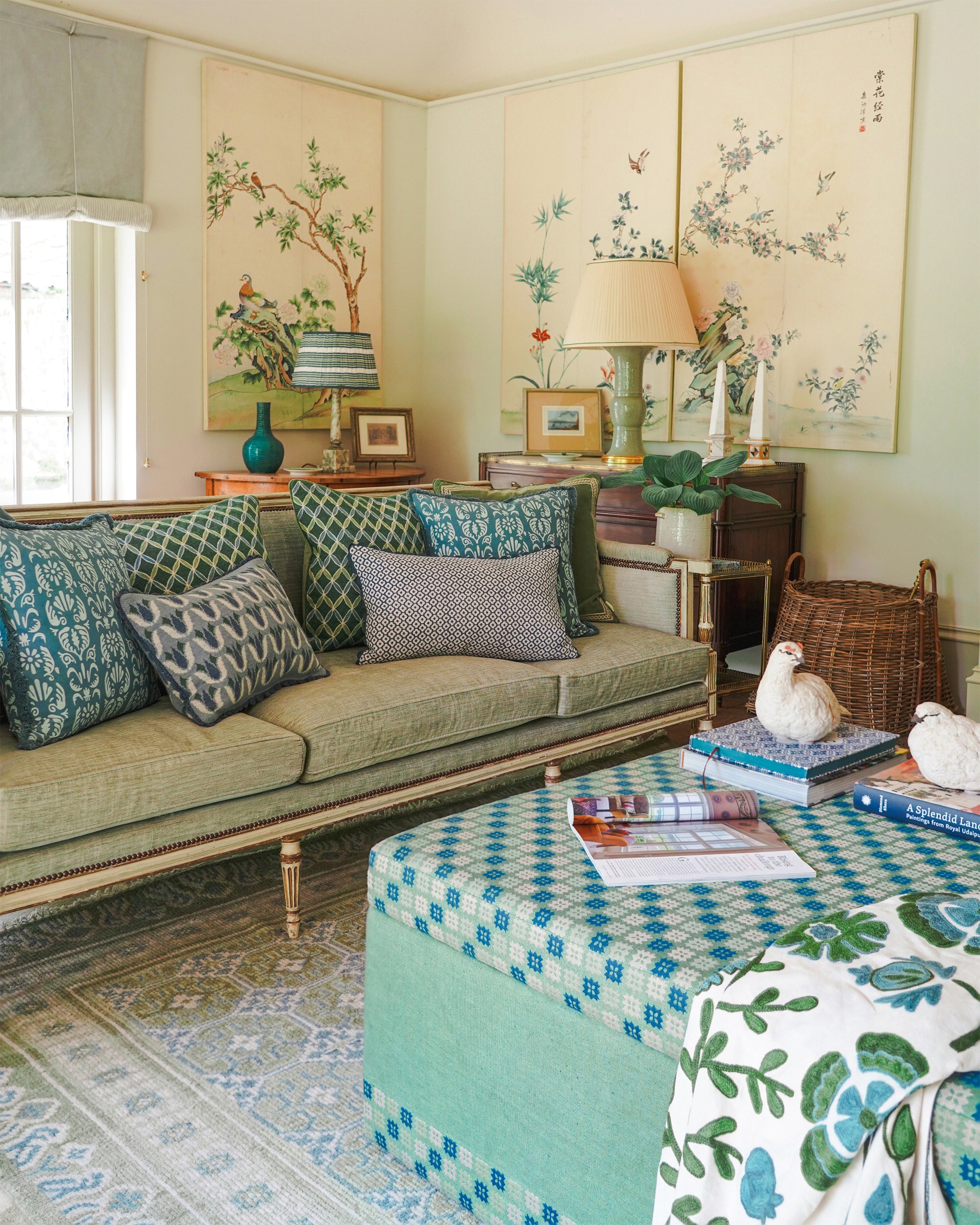 A styled living room featuring a sage green sofa with blue and gray patterned pillows, a matching blue and green patterned ottoman coffee table, and a blue-and-white floral throw blanket. The room is decorated with traditional-style botanical wall hangings, a patterned area rug, and a sideboard with lamps and decorative items.