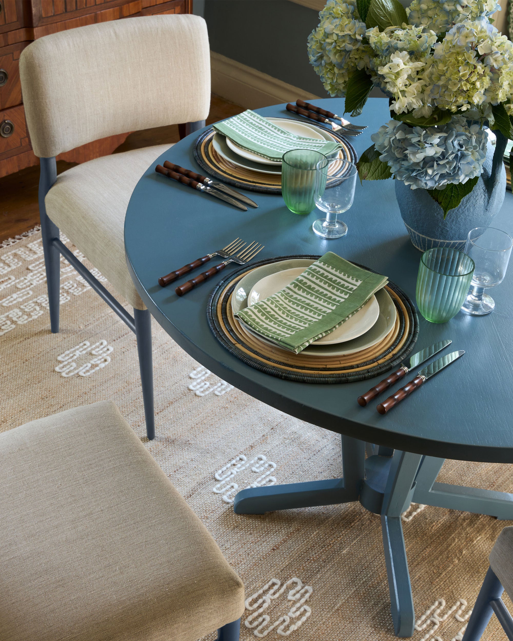Blue tablscape featuring Wensum pedestal table, blue rattan placemats, green bel napkin and cucito rug
