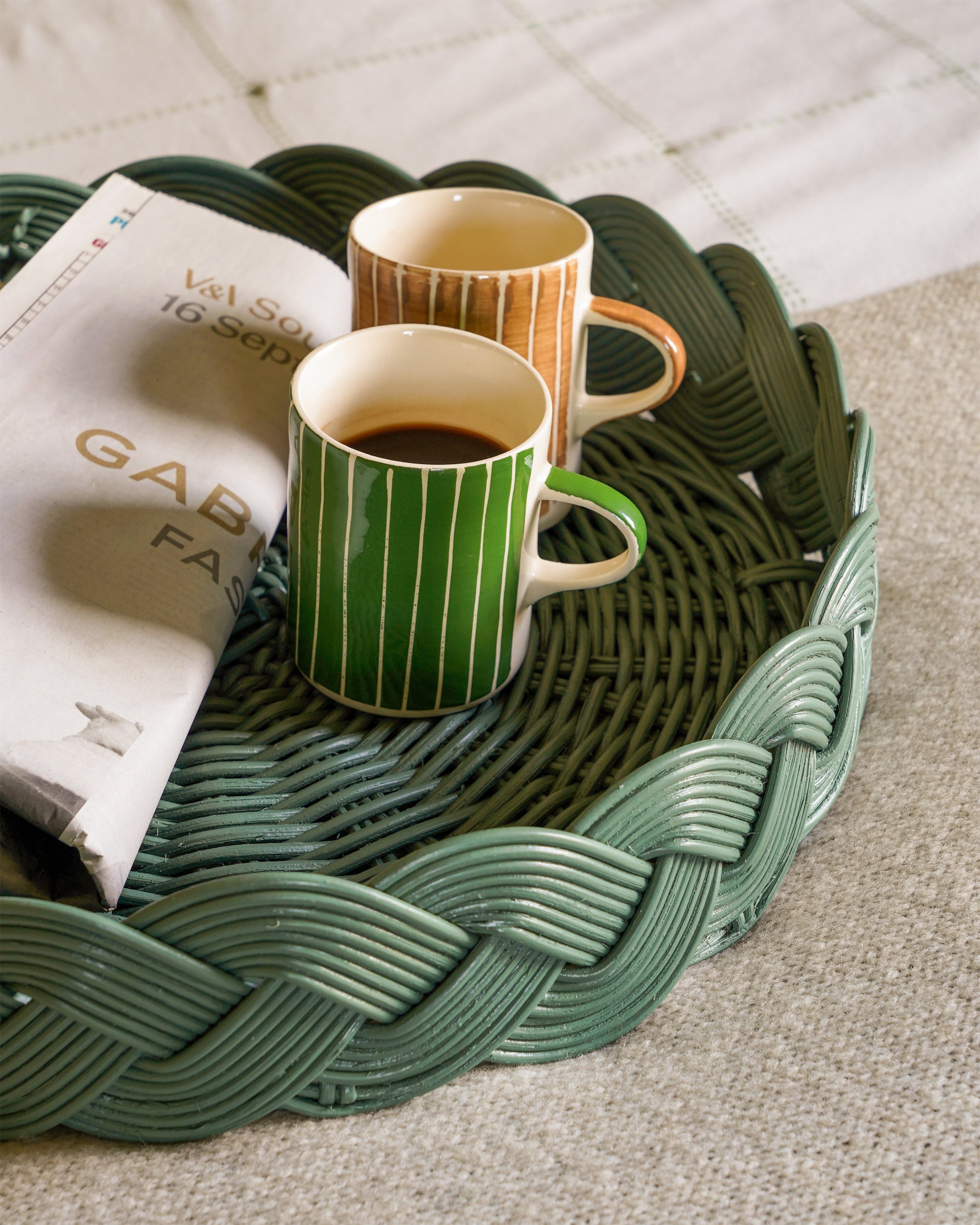 Two mugs on a woven green tray with a newspaper underneath.