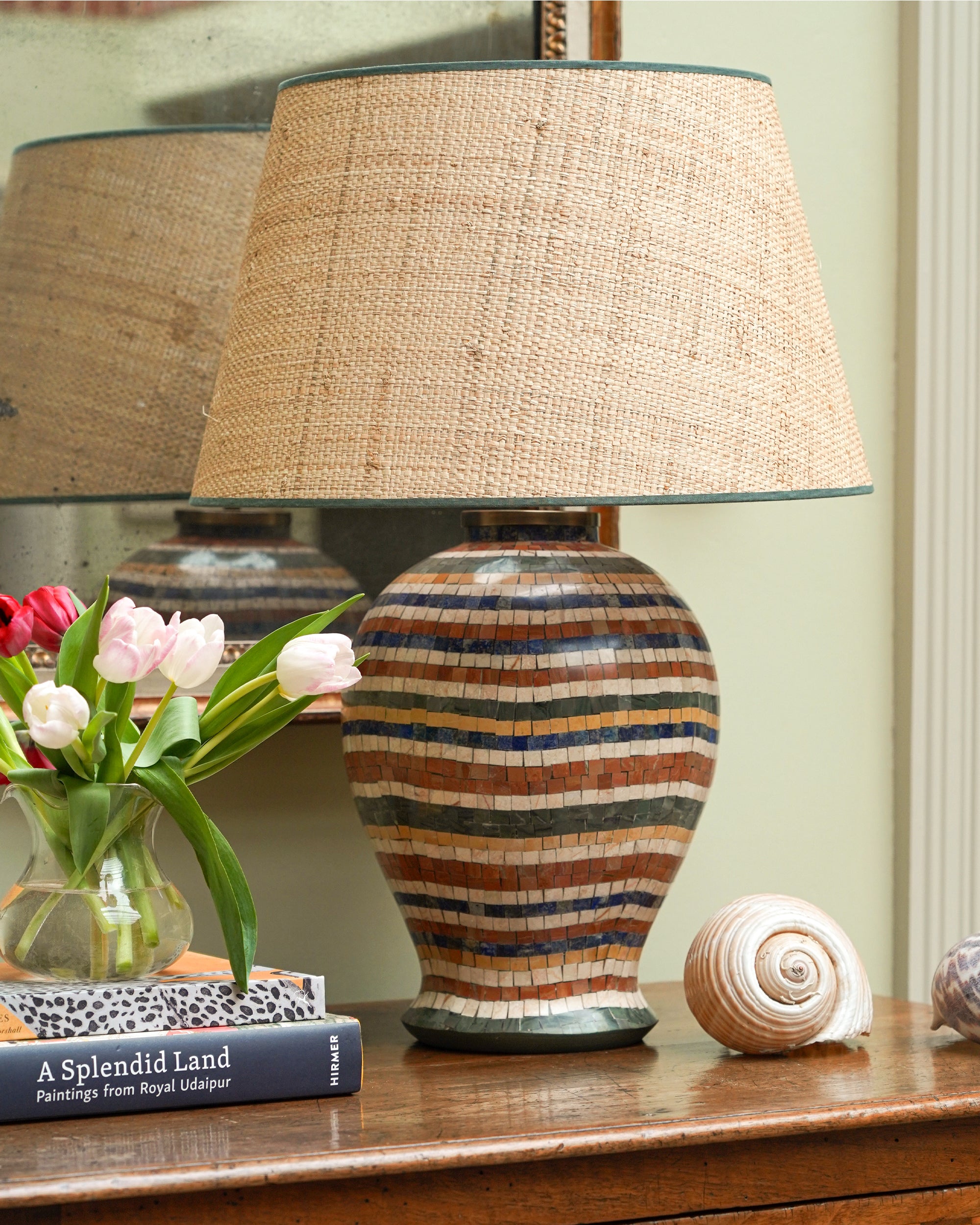 A mosaic lamp with a striped ceramic base and a burlap shade, placed on a wooden surface, with a seashell and a book with tulips in the background.