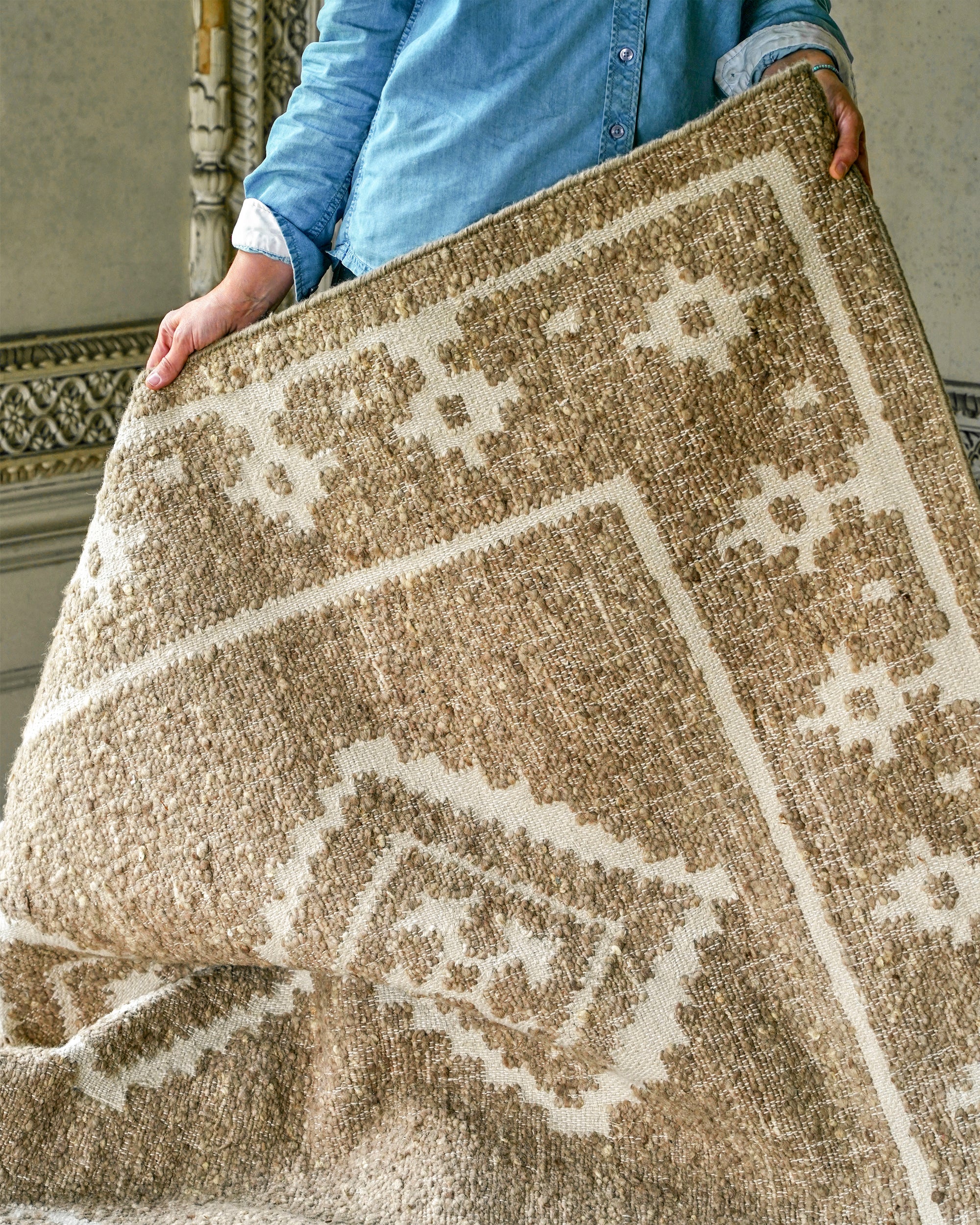 Person holding a patterned beige rug indoors