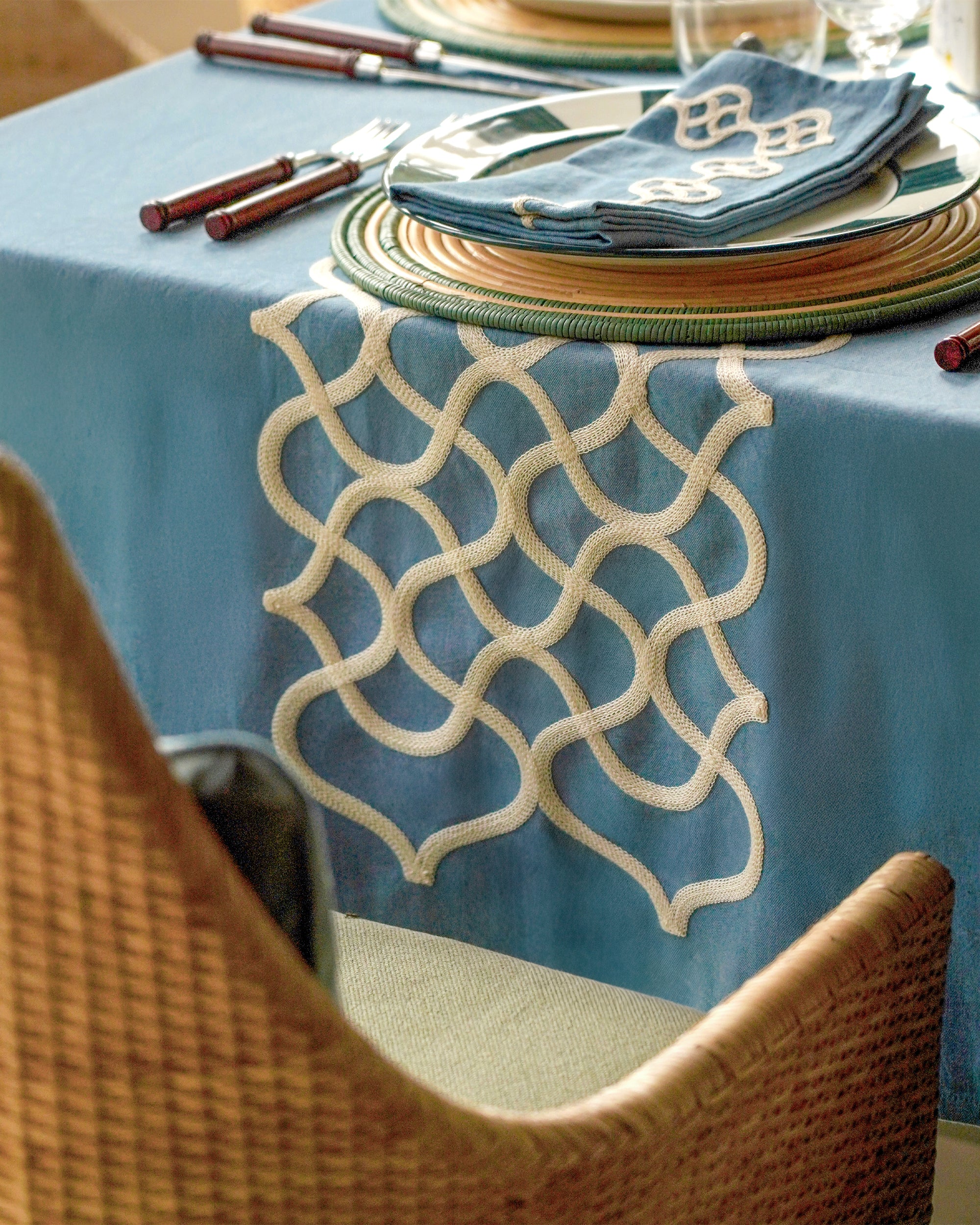 Table setting with blue tablecloth, patterned placemat, and cutlery.