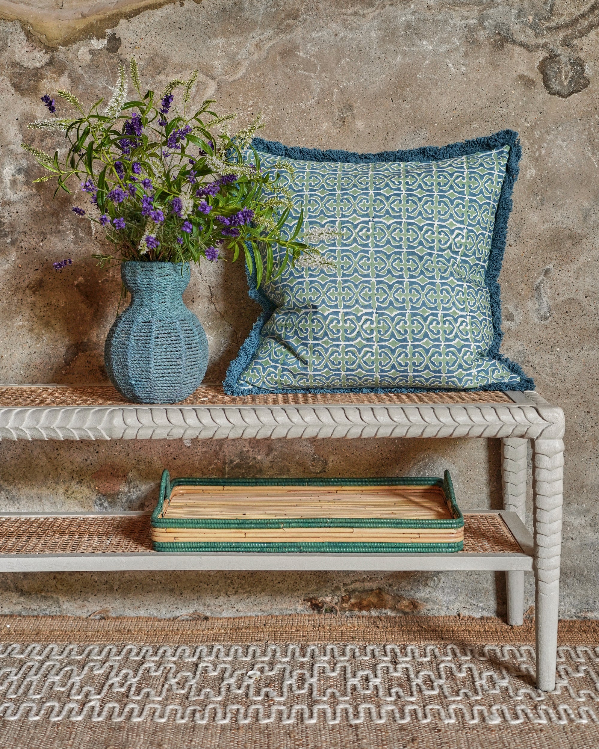 Decorative pillow on a bench with a vase of flowers and a wooden tray against a textured wall.