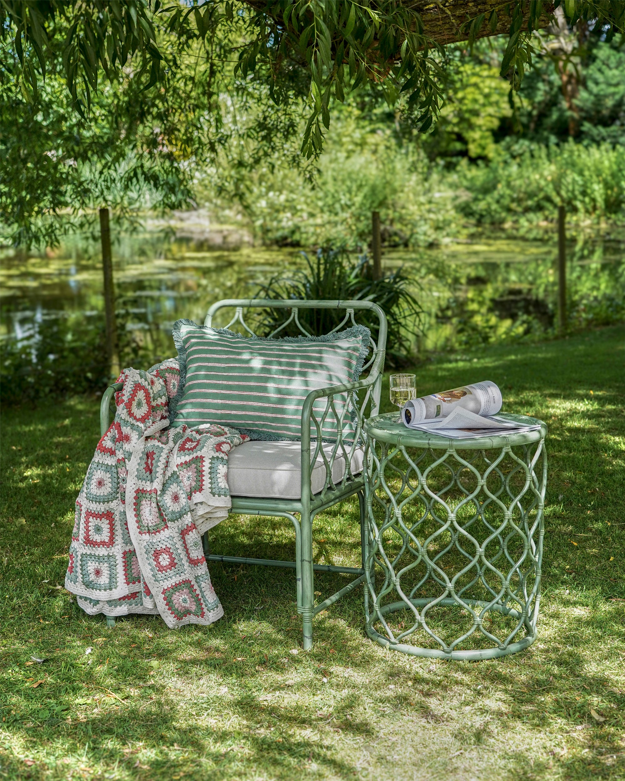 Garden setting with a chair, table, and blanket under a tree by a pond.