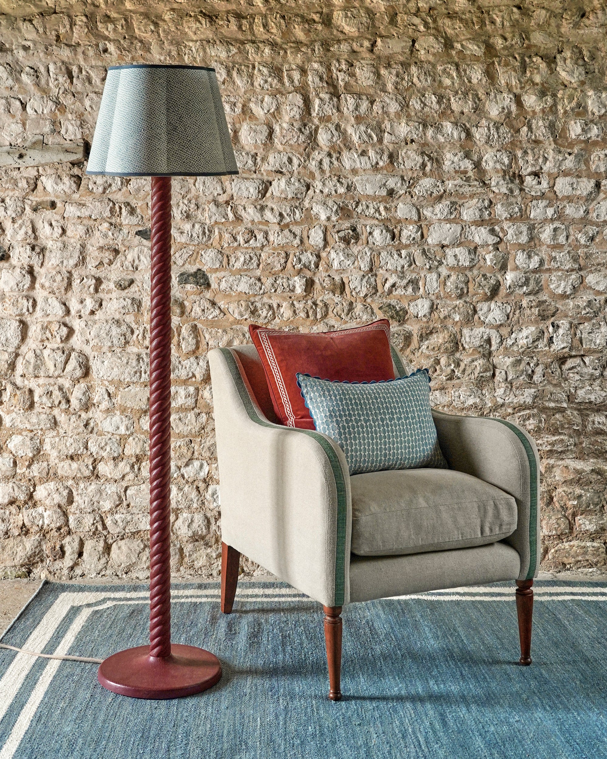 Chair with cushions next to a floor lamp against a stone wall.