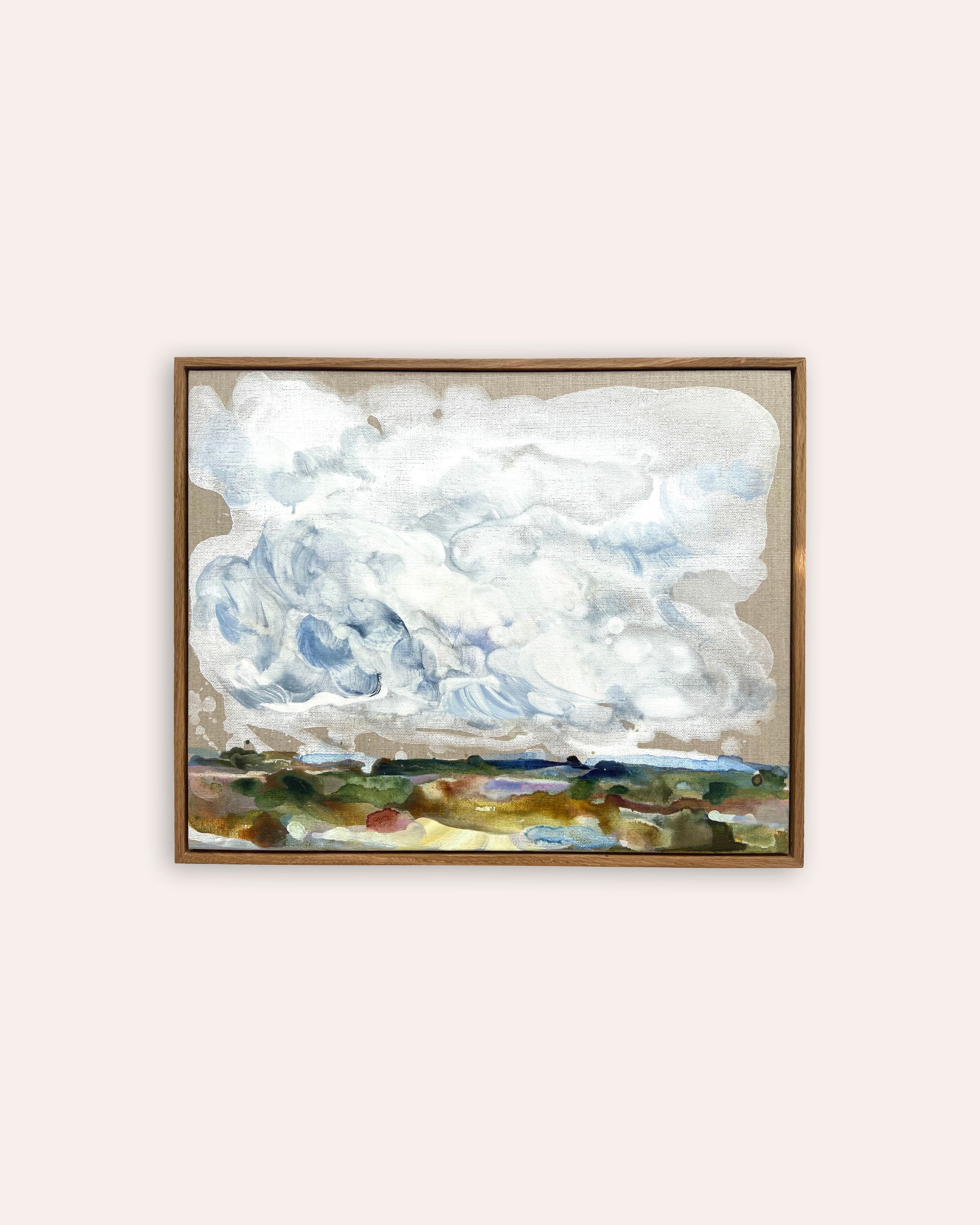A framed landscape painting featuring dramatic white and blue clouds over a green and brown field, displayed on a light beige wall.
