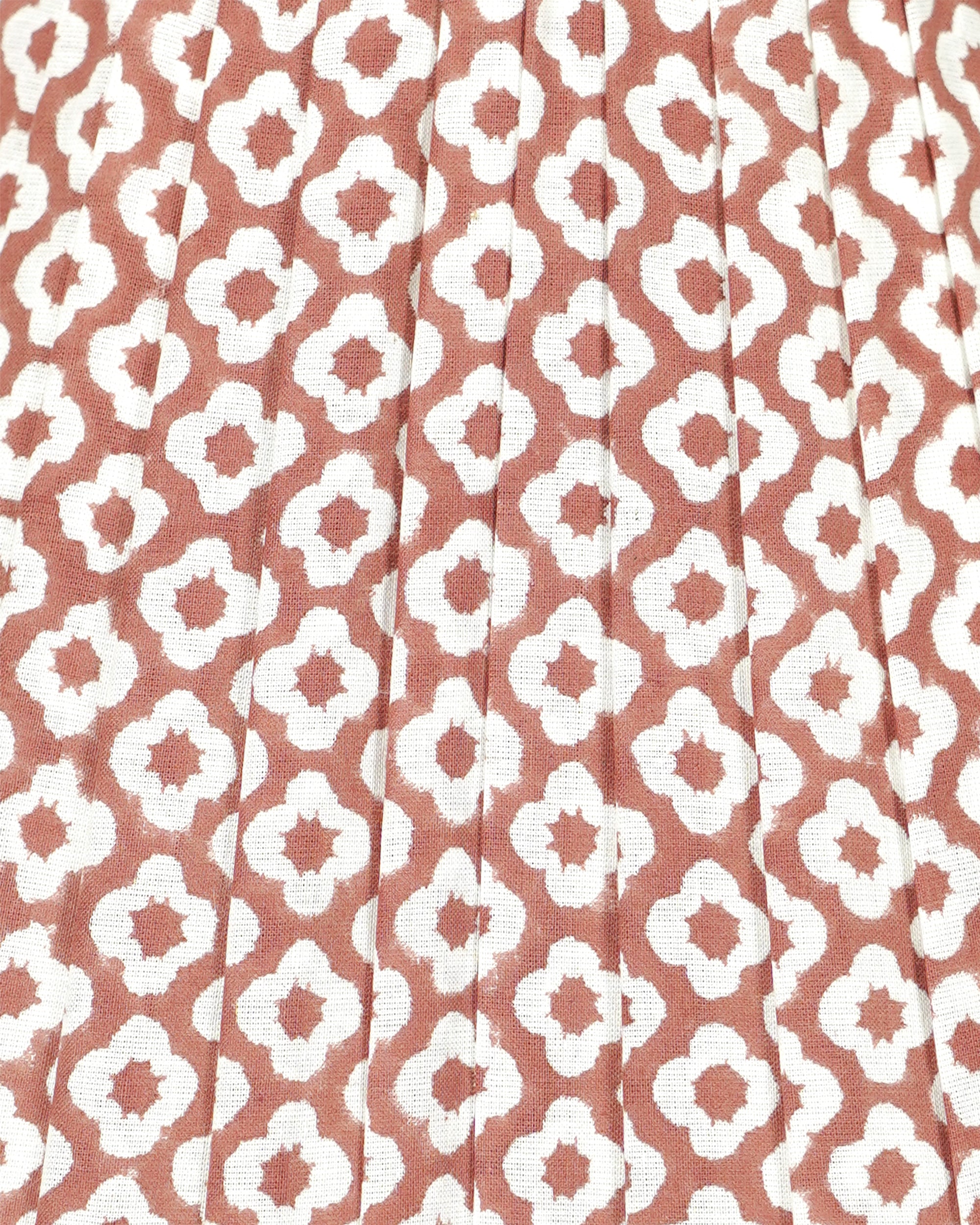 A close-up of a pleated fabric with a repeating white ikat-style geometric pattern on a muted terracotta or dusty rose background.