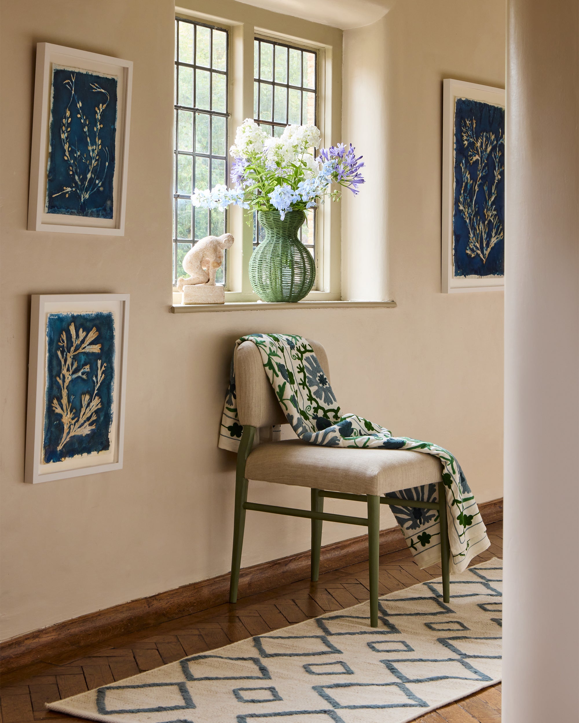 Blue Wensum chair in front of a window with a vase of flowers, framed artwork on the wall.