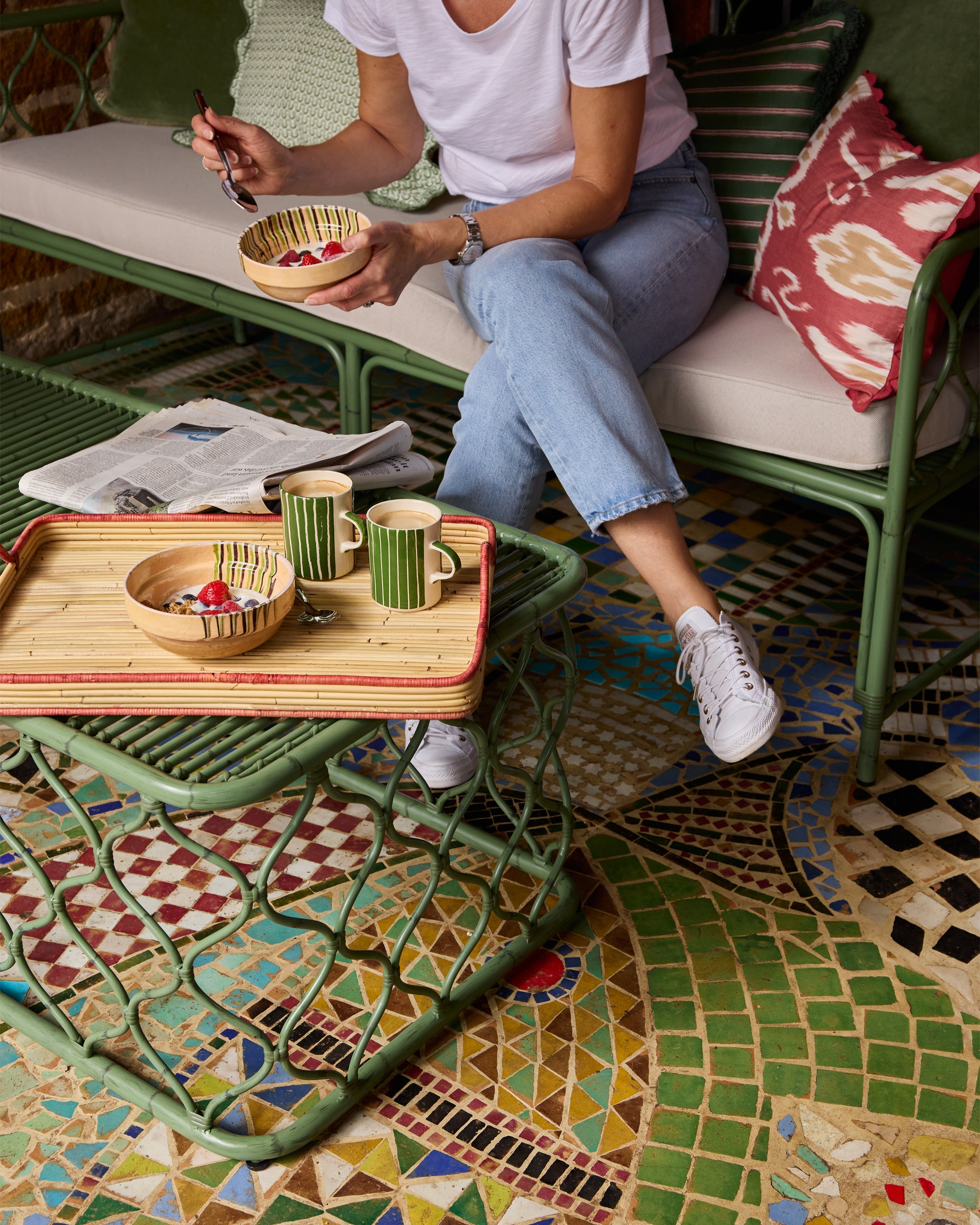 Person sitting on a the green Curato sofa with a red Rattan tray of food and drinks on a mosaic-tiled floor.