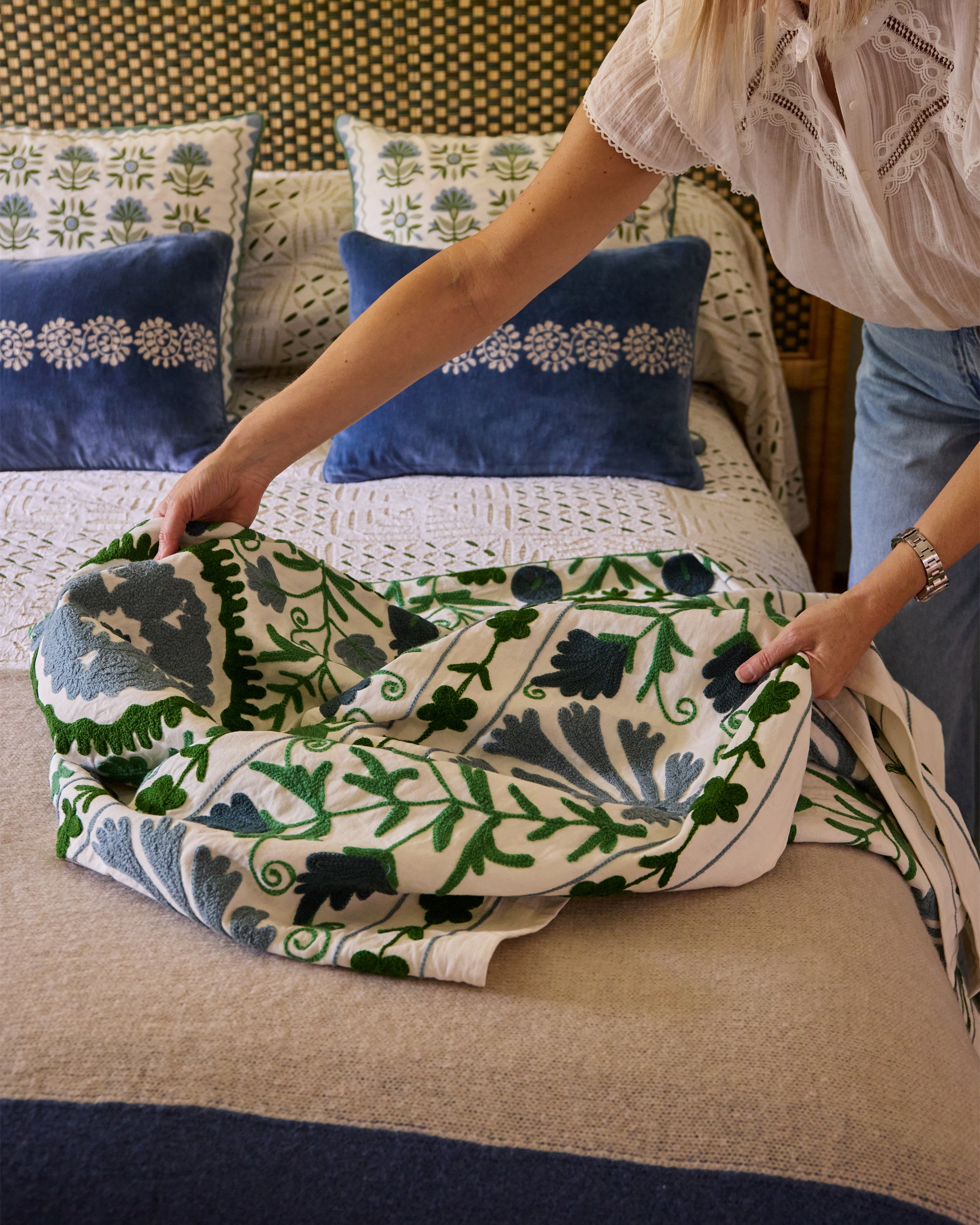 Person holding the blue and green Dumaloq Suzani throw on a bed with decorative cushions in the background