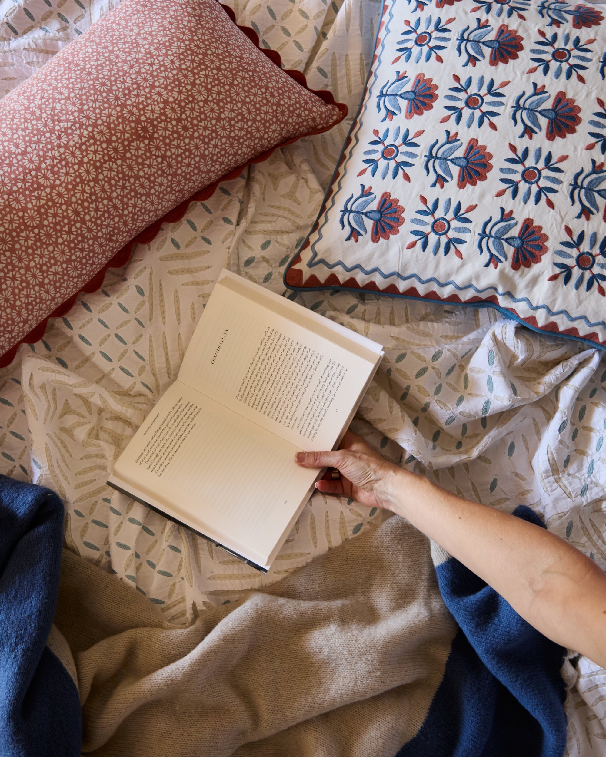 Relaxed flat lay of someone reading a book amongst Birdie Fortescue's pink and blue spring summer cushions