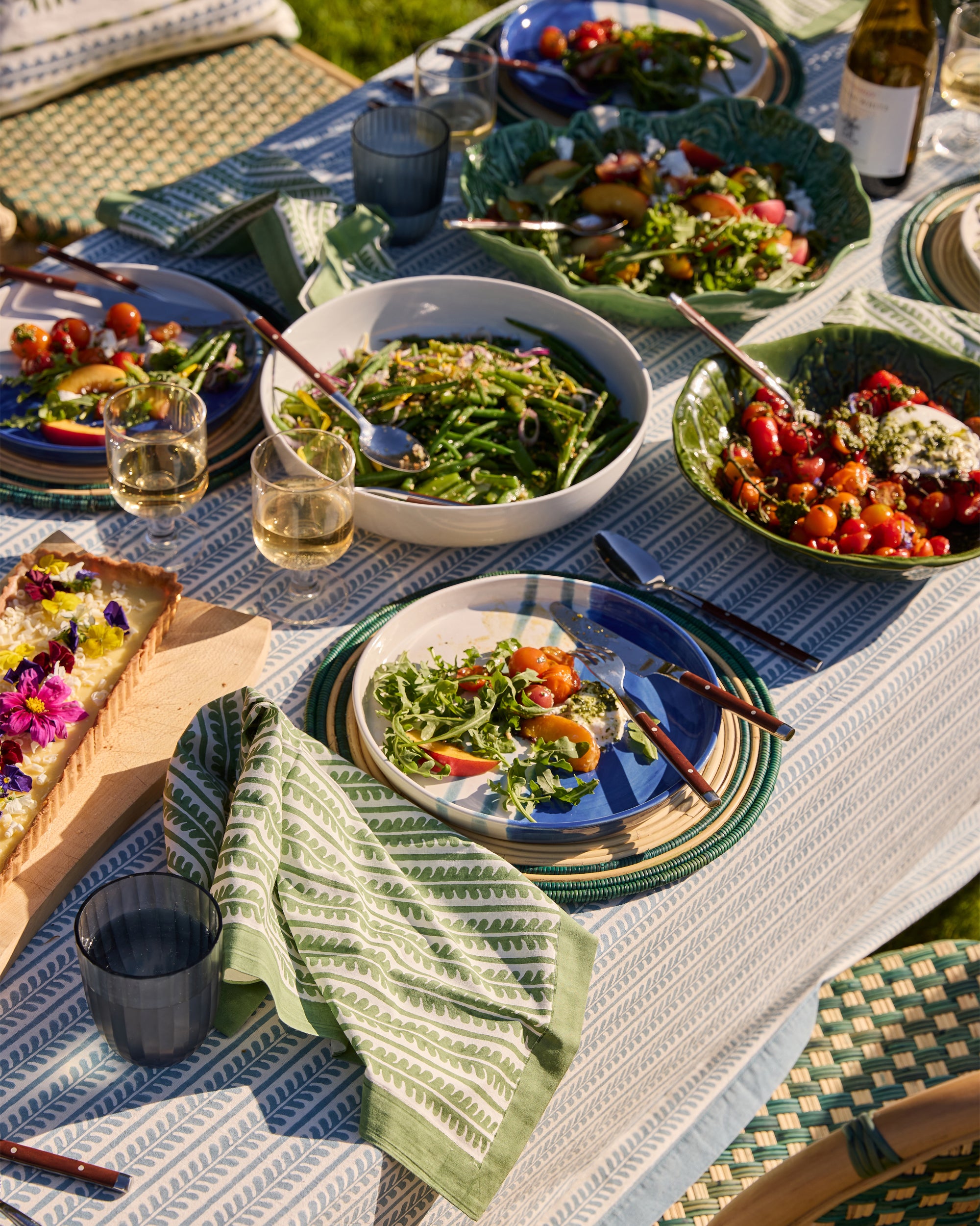 Outdoor dining table set with various dishes, including salads and a pie, on a patterned tablecloth.