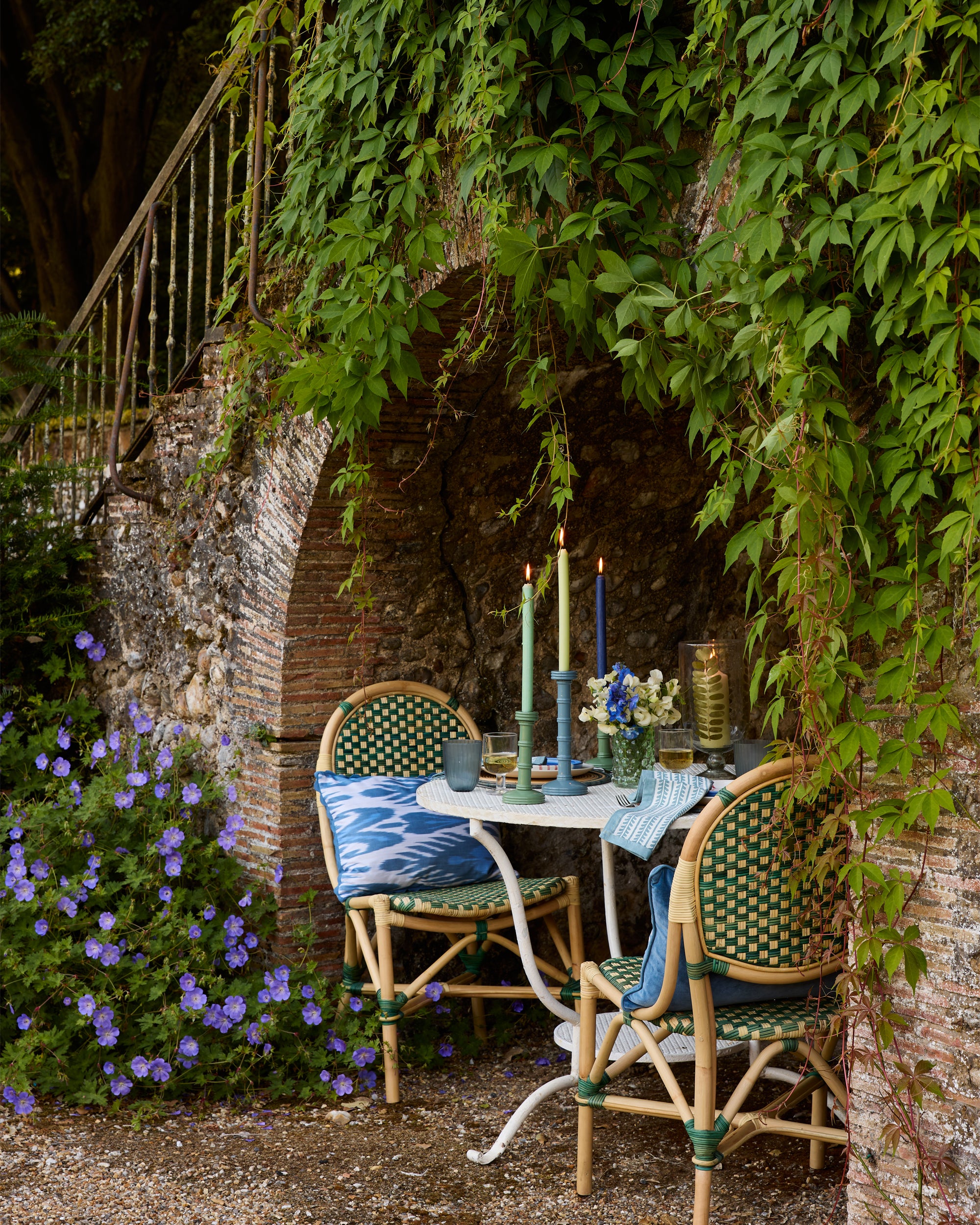 Outdoor dining setup with chairs, table, and candles under a stone archway with greenery.