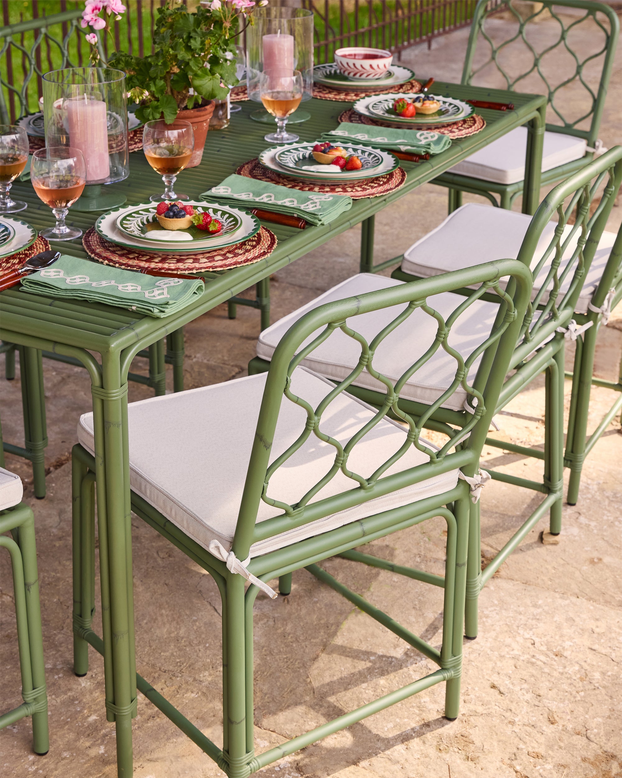 Outdoor dining table set with green chairs and white cushions, featuring plates and glasses on a stone patio.