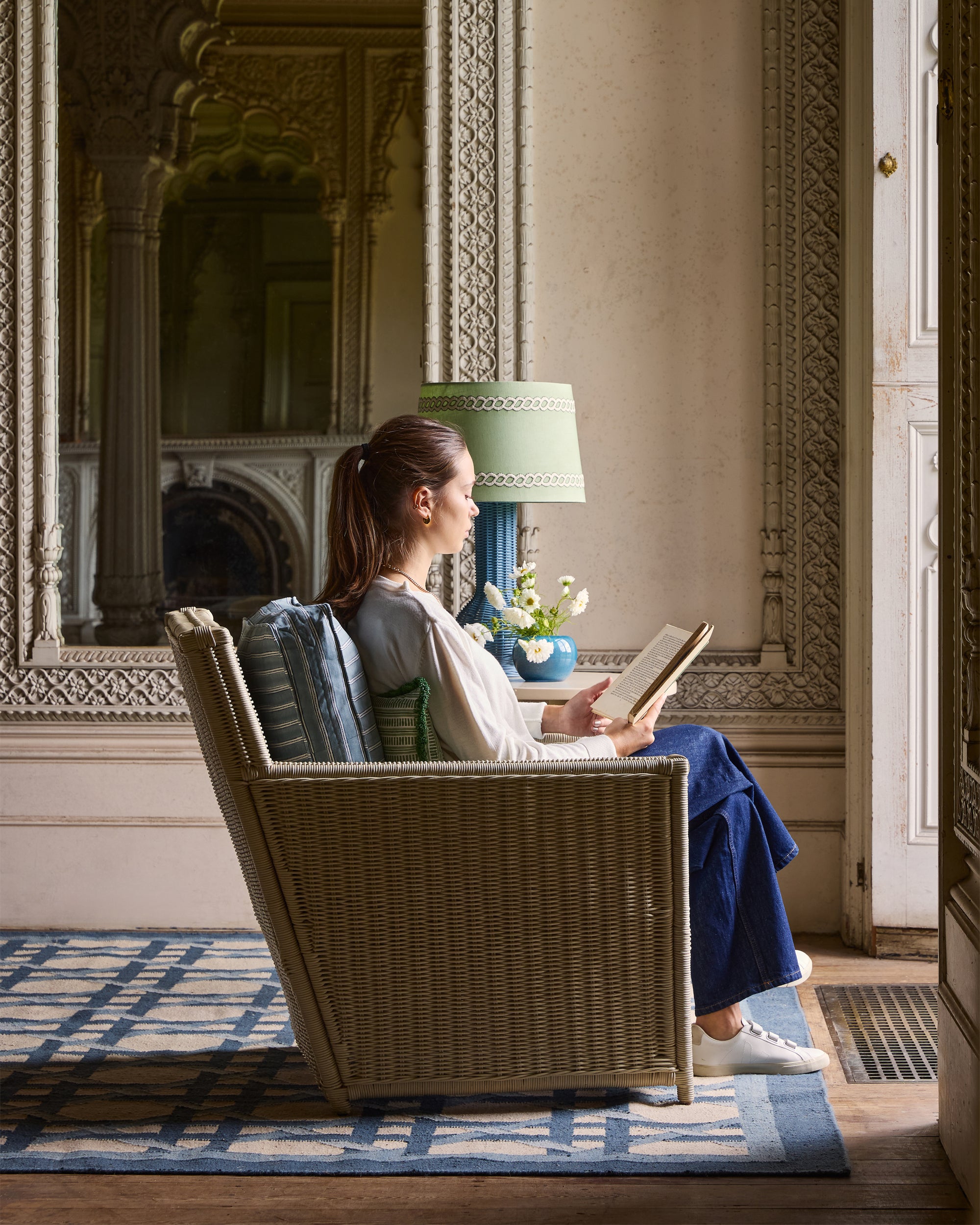 Woman reading a book in a ornately decorated room with a lamp and decorative elements.