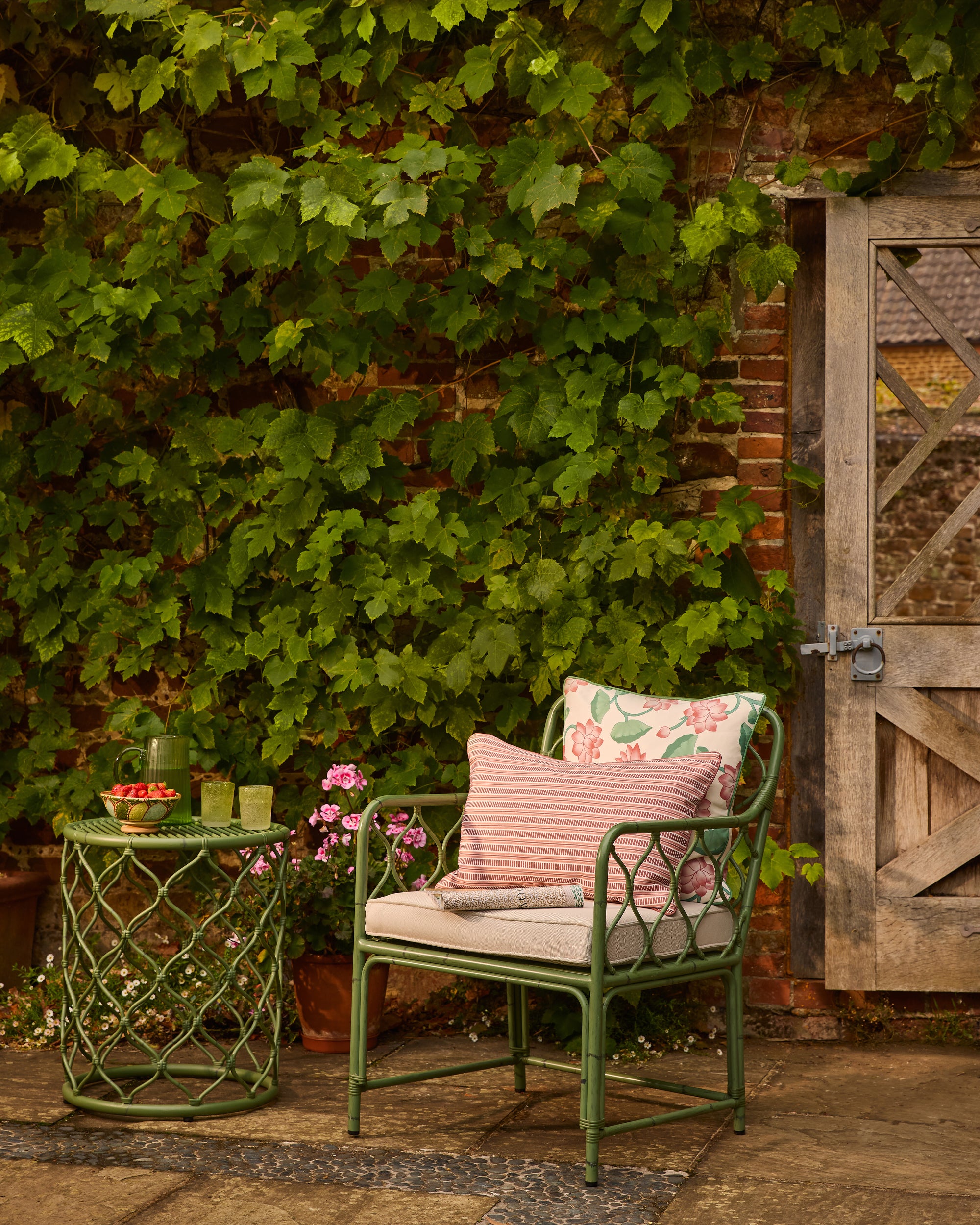 Green metal chair with floral and pink cushions against a brick wall with greenery