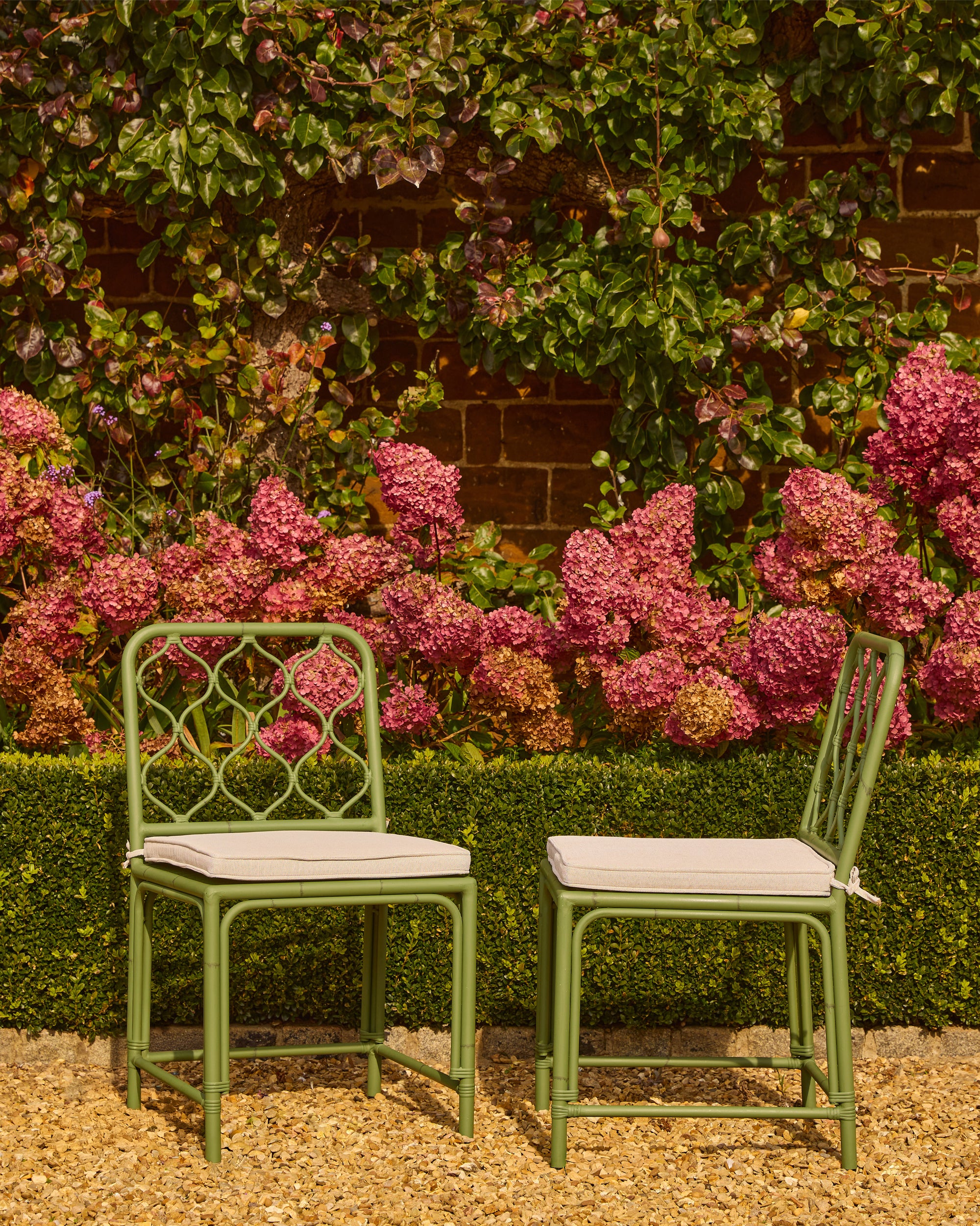Two green garden chairs with white cushions in front of a flowering bush and brick wall.