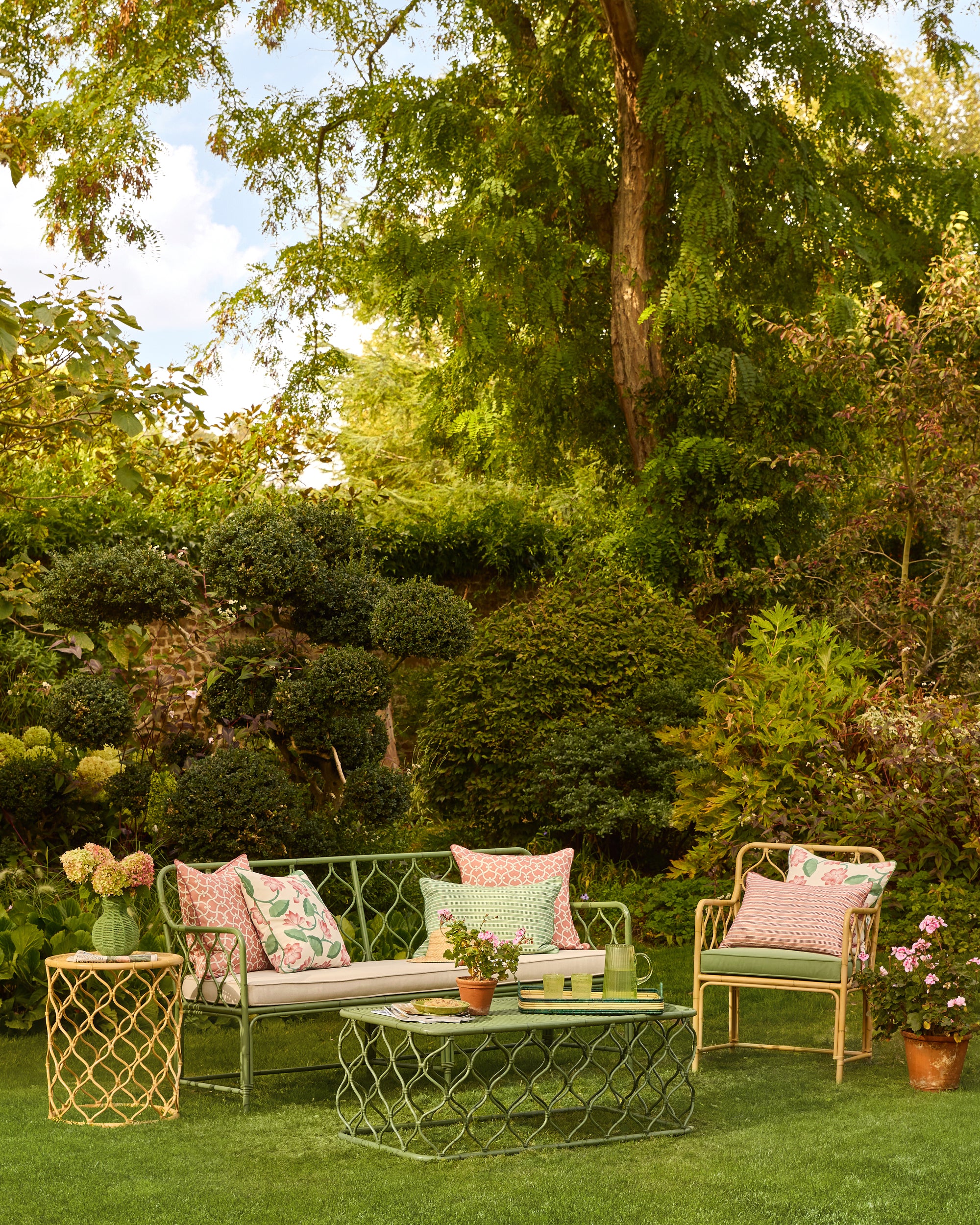 Outdoor garden setting with green furniture and decorative pillows surrounded by trees and plants.