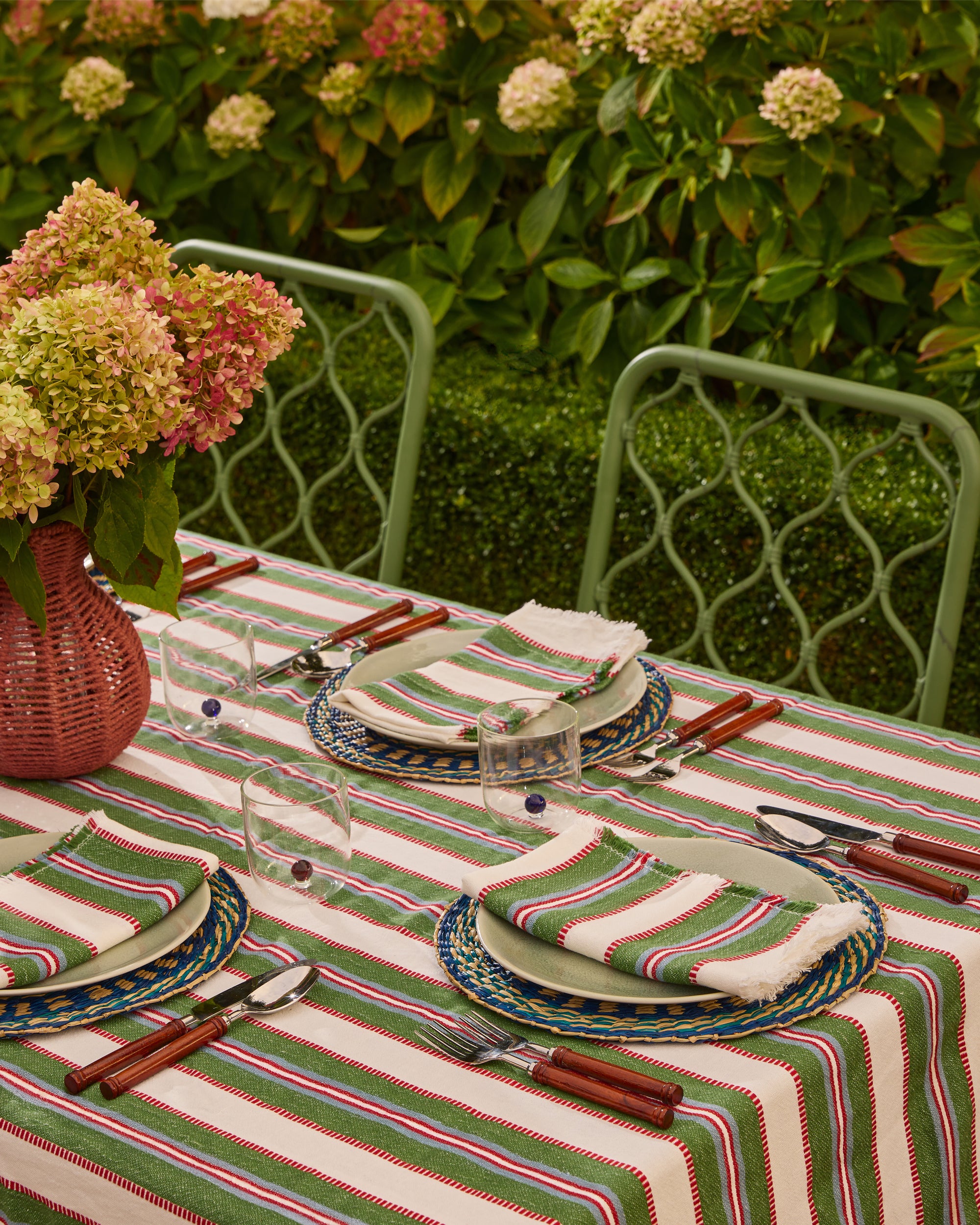 Outdoor table setting with striped tablecloth, floral arrangements, and green chairs.