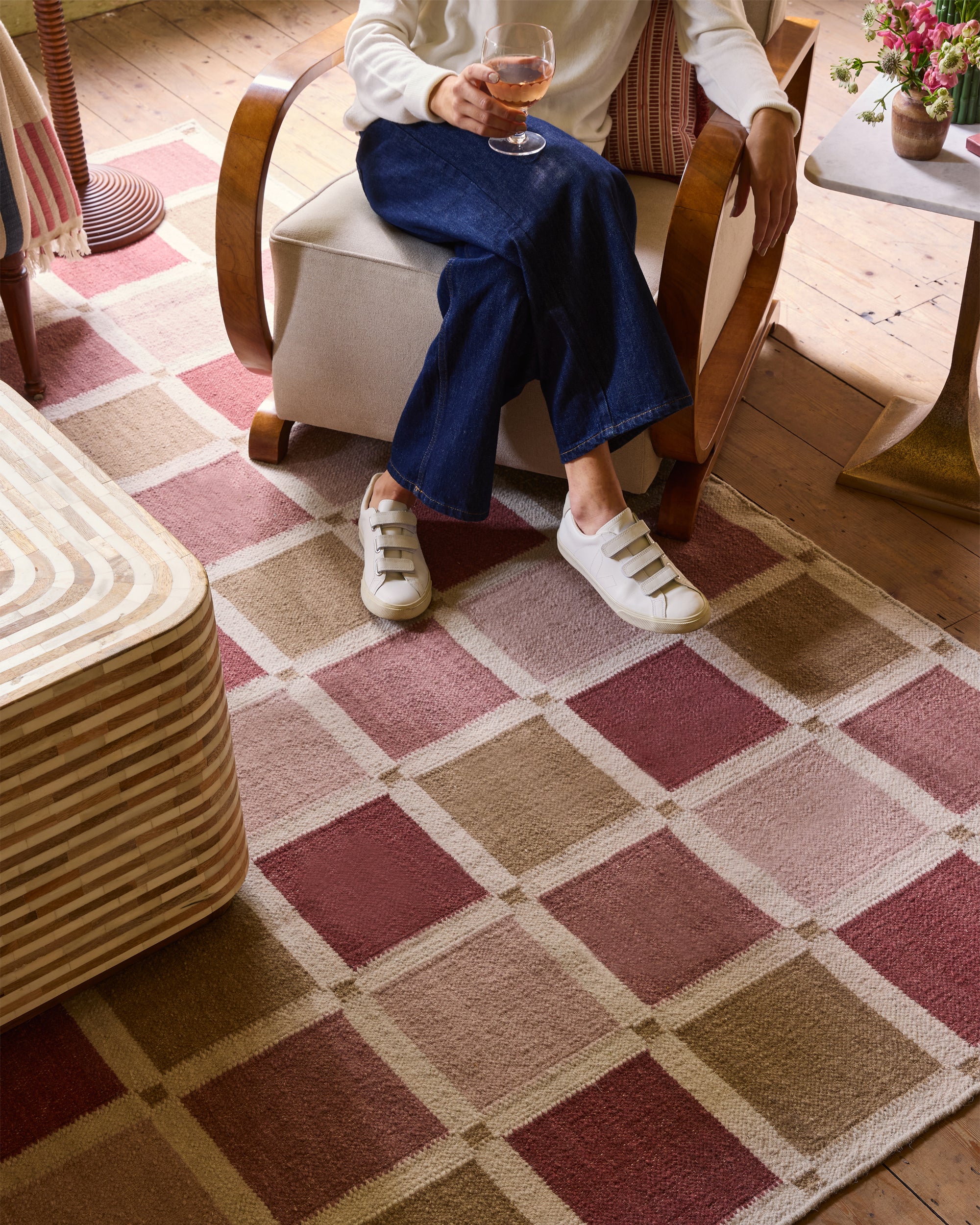 Person sitting on a chair with a glass of wine, wearing white sneakers and blue jeans, on a checkered rug.