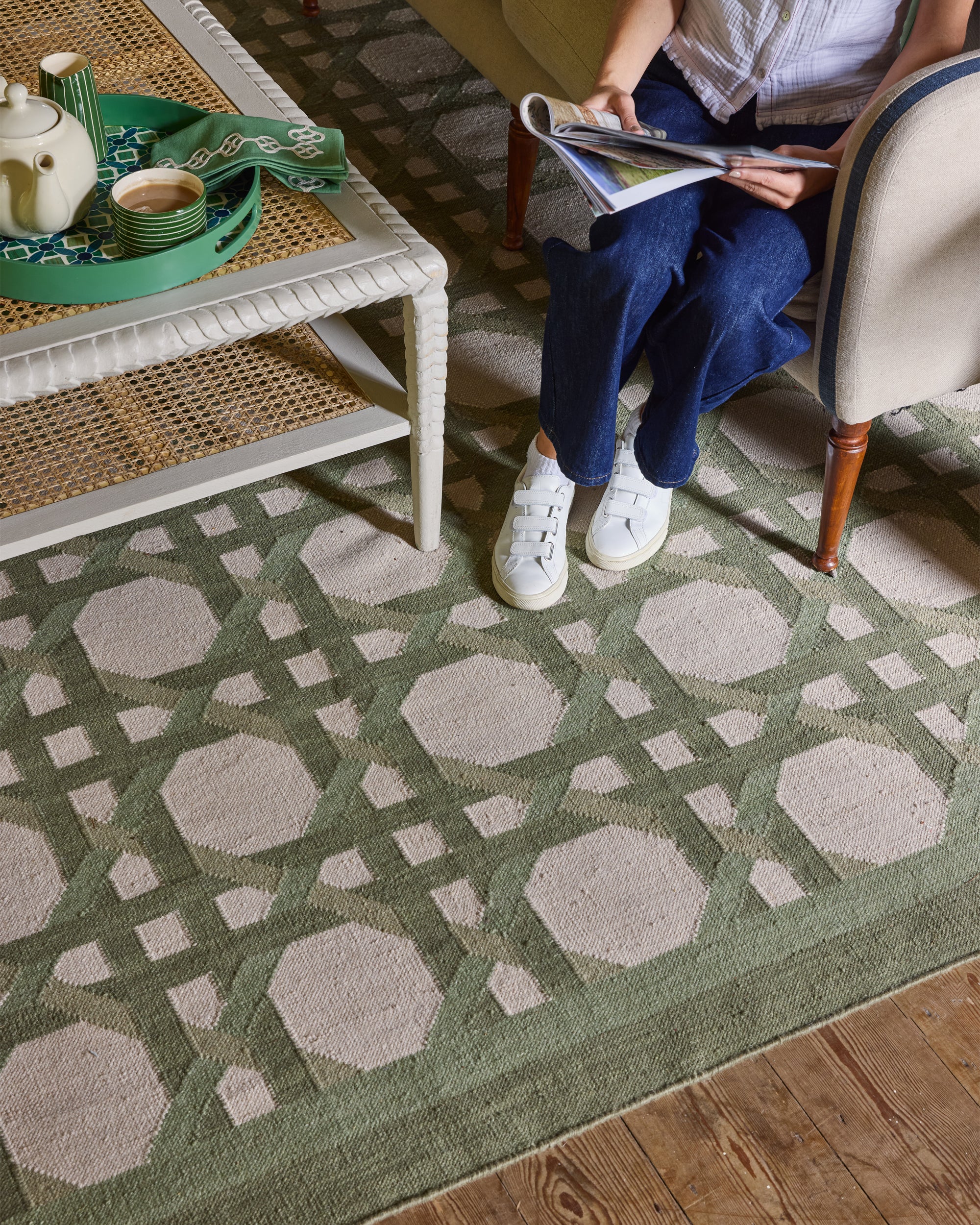 Person sitting on a patterned green rug in a room with a table and chair.