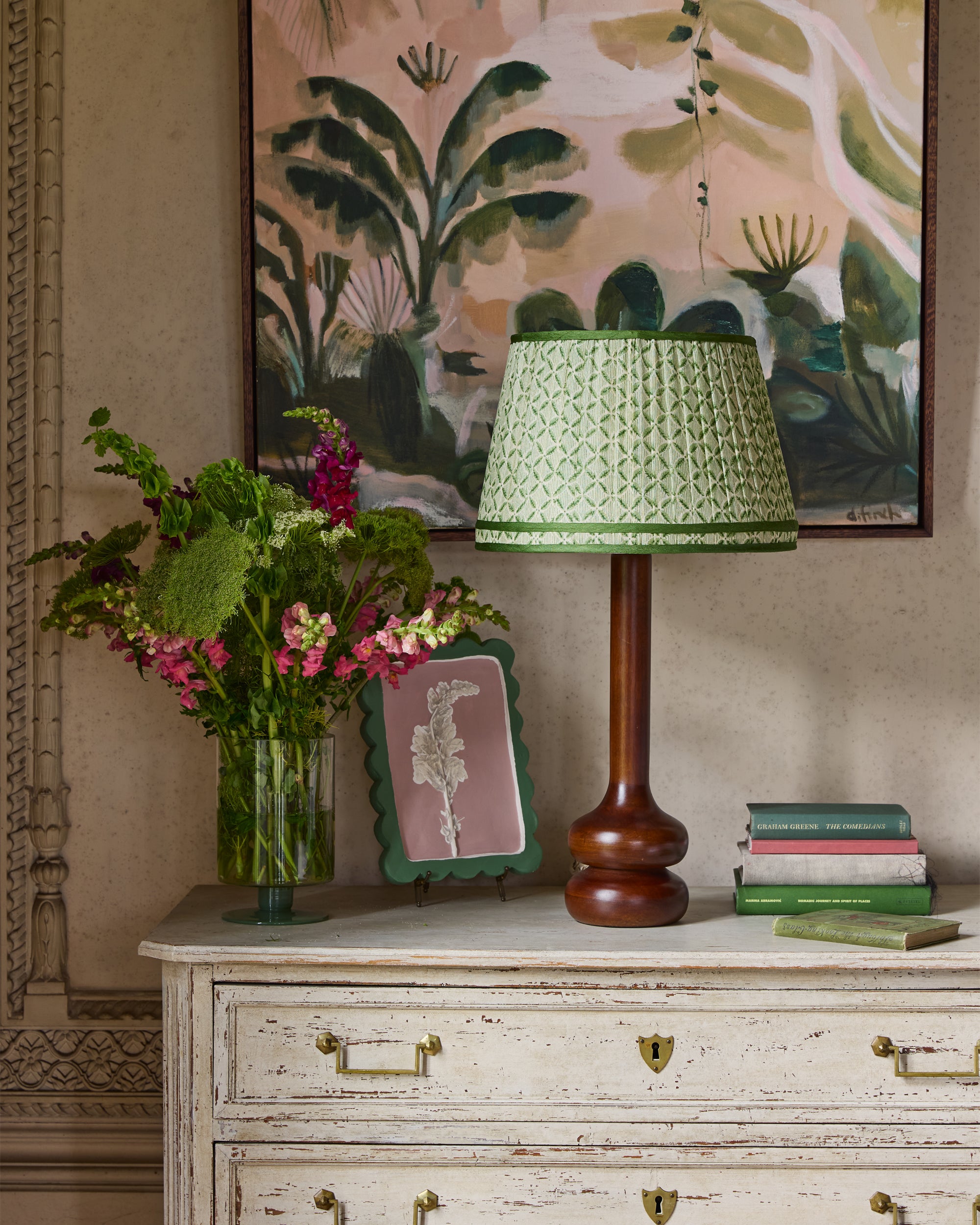 Decorative lamp with green shade on a wooden cabinet with floral arrangement and books.