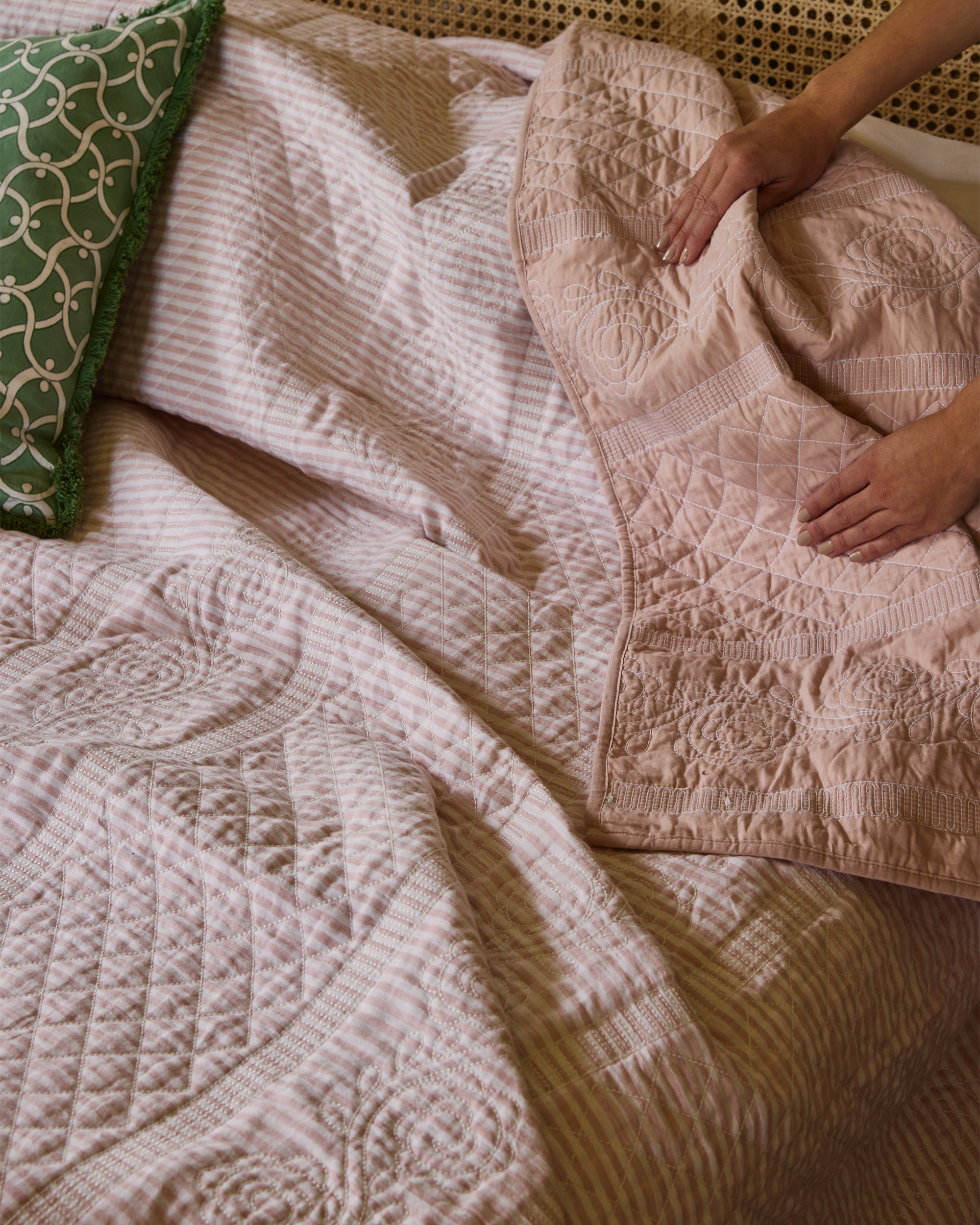 Person arranging a textured pink blanket on a bed with a green pillow.