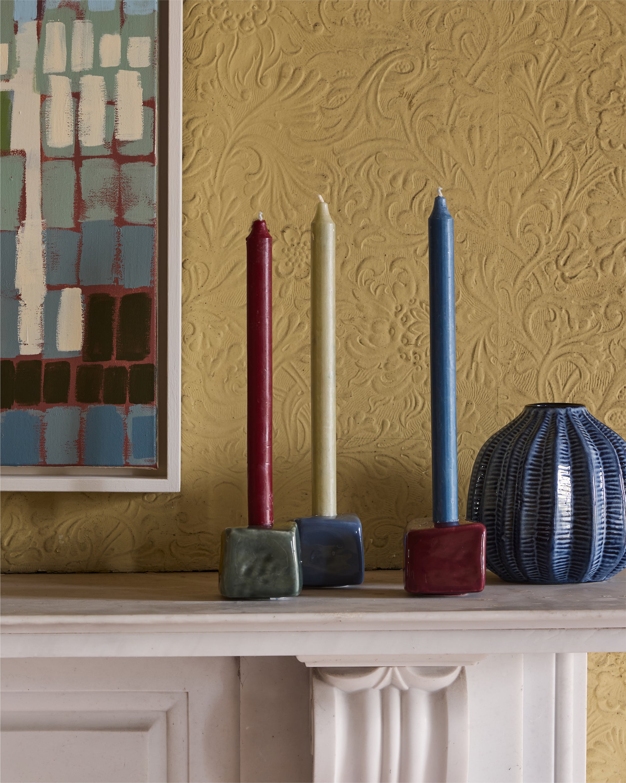 Decorative candles and vases on a mantelpiece with a textured gold wall and abstract painting in the background.