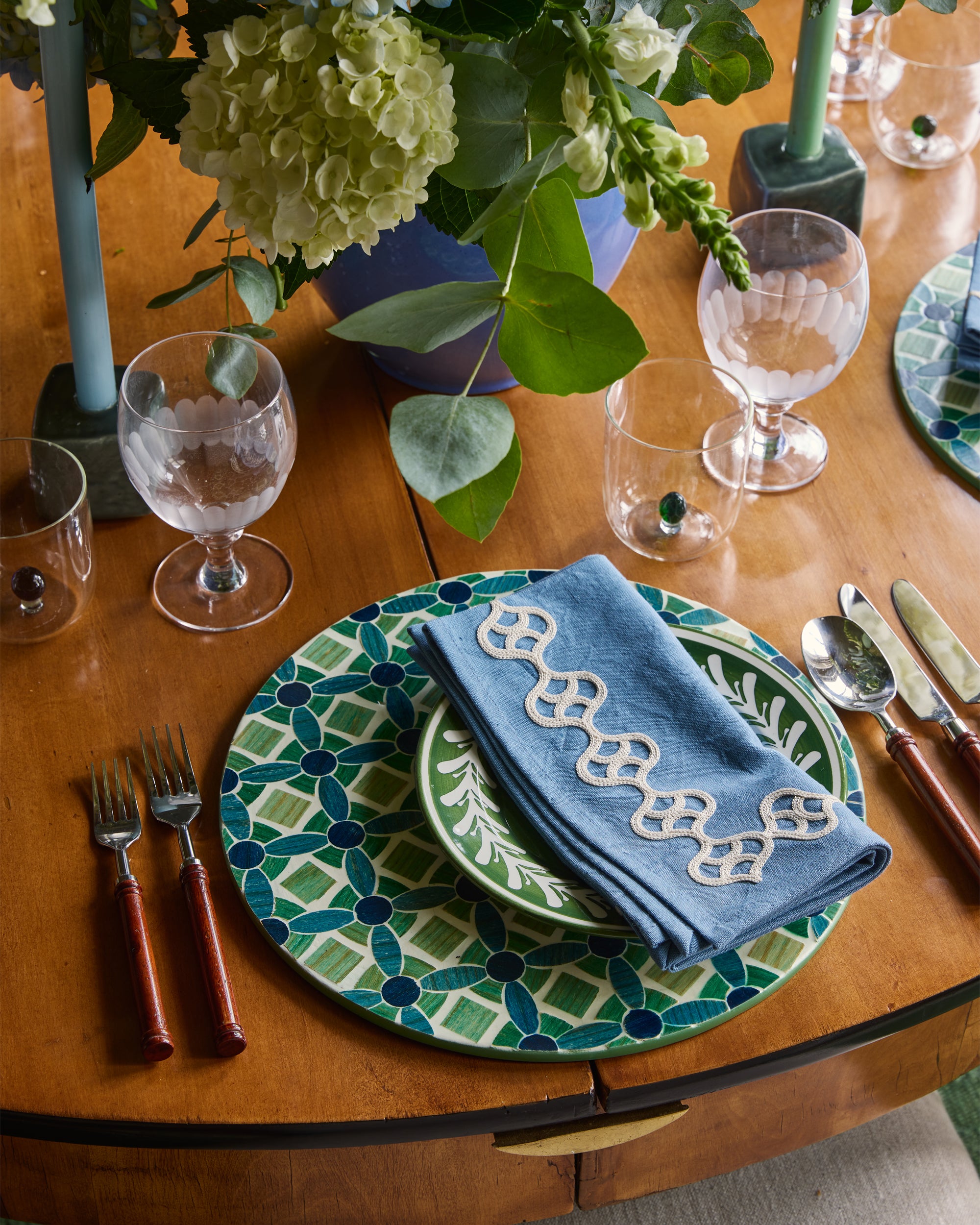 Dining table setting with green and blue patterned plates, silverware, and a floral centerpiece.