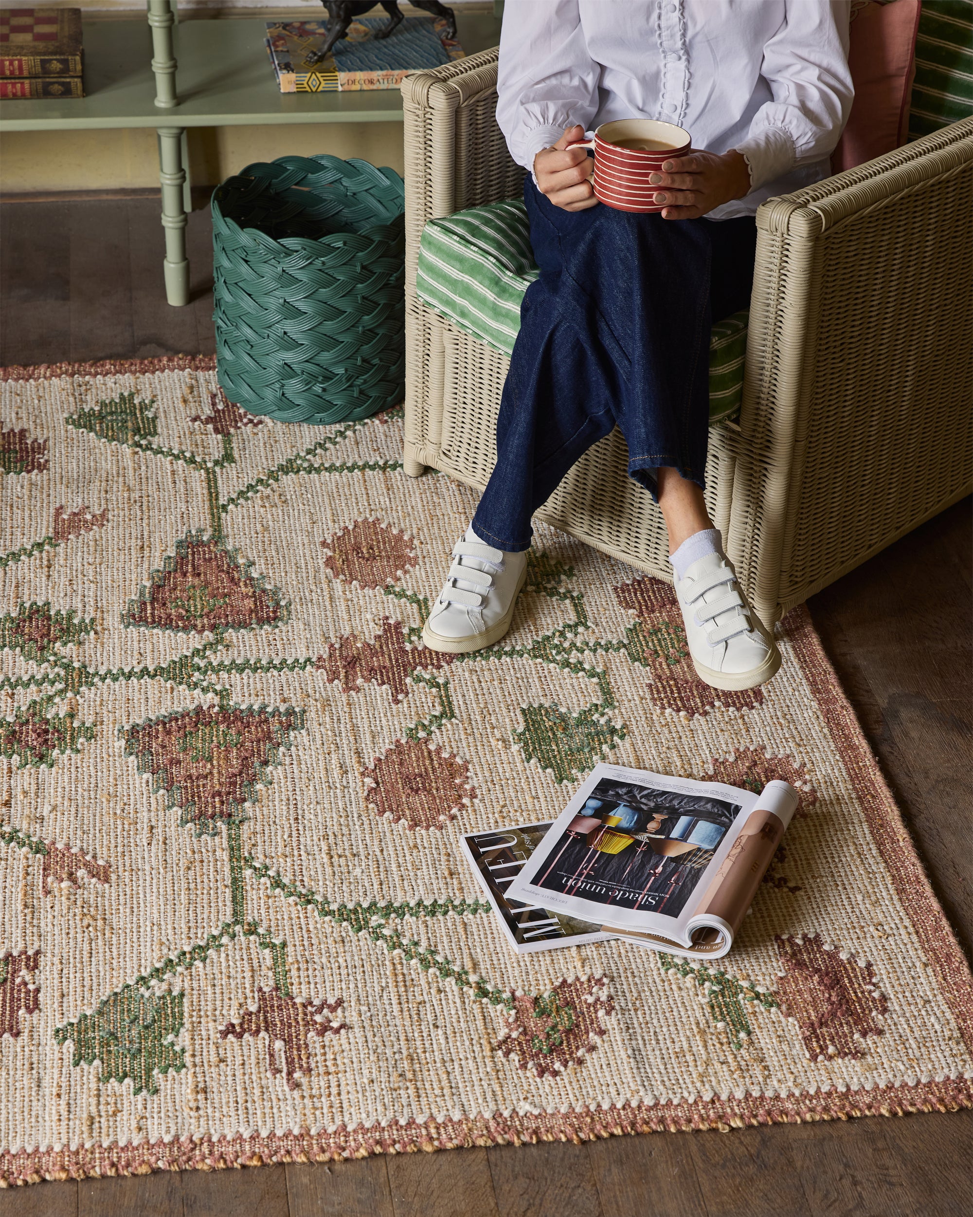 Person sitting on a chair holding a cup, with a patterned rug and magazine on the floor.