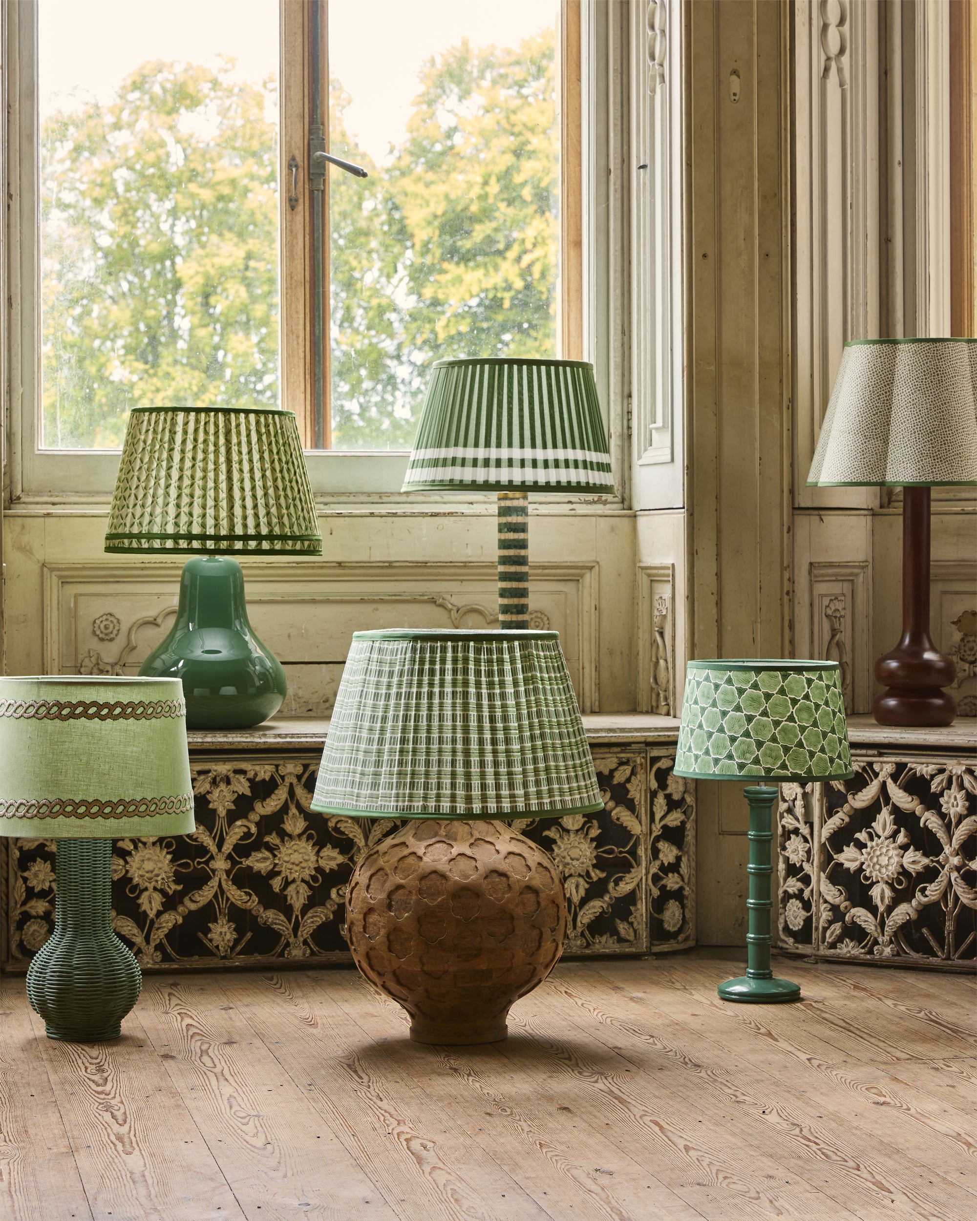 Various lamps with green and brown shades in a room with wooden floors and large windows.