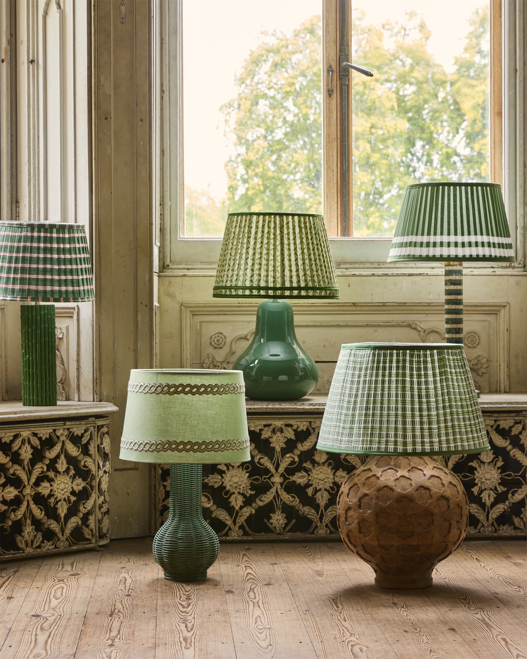 Collection of green lamps with patterned shades in a room with wooden floor and large window.