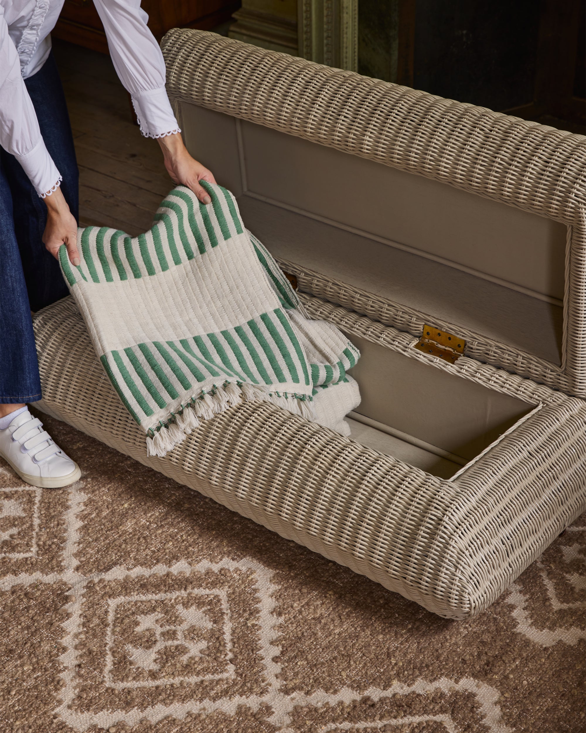 Person placing a green and white striped cushion into an open wicker storage bench.