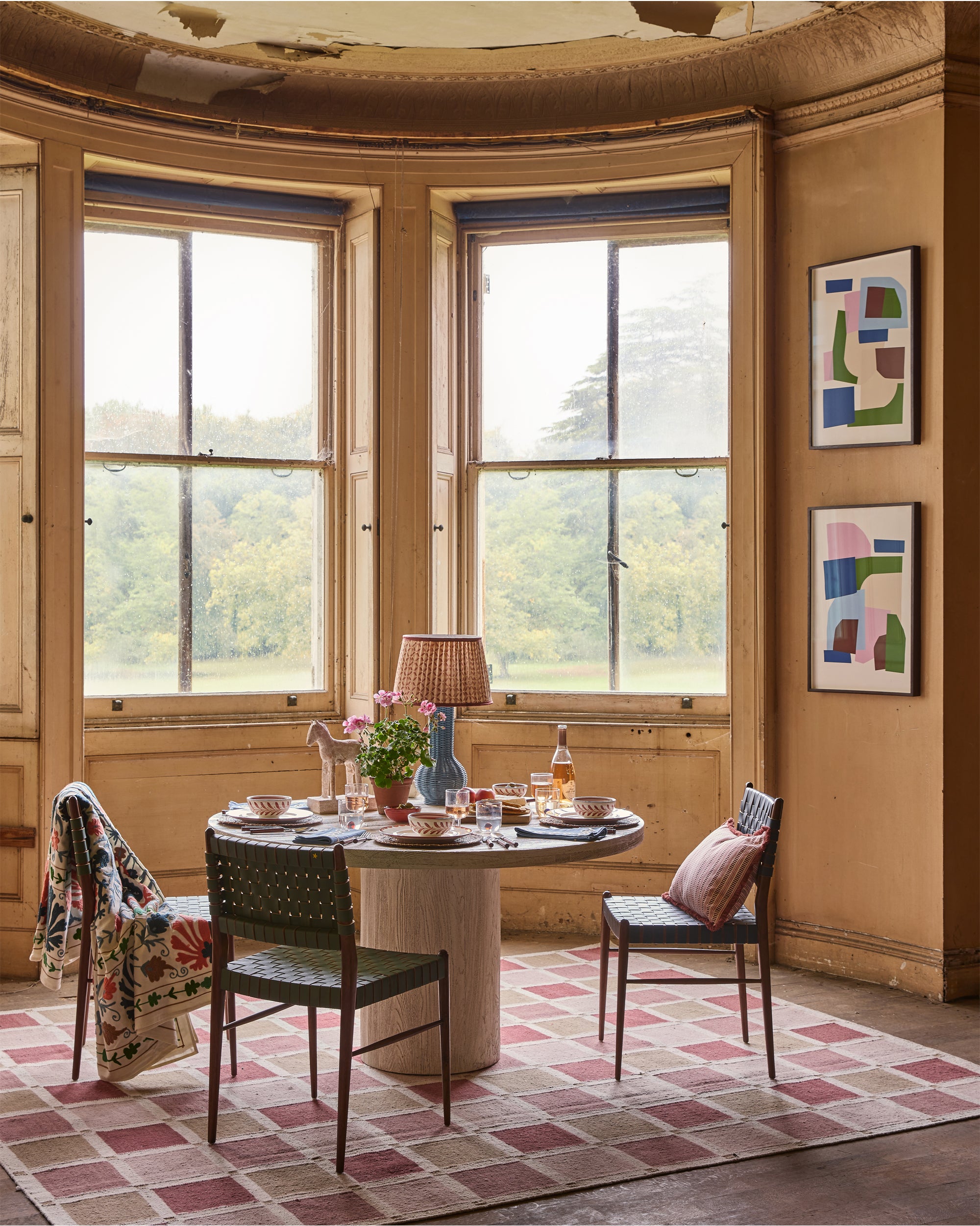 Dining room with round table, chairs, and large windows overlooking greenery.