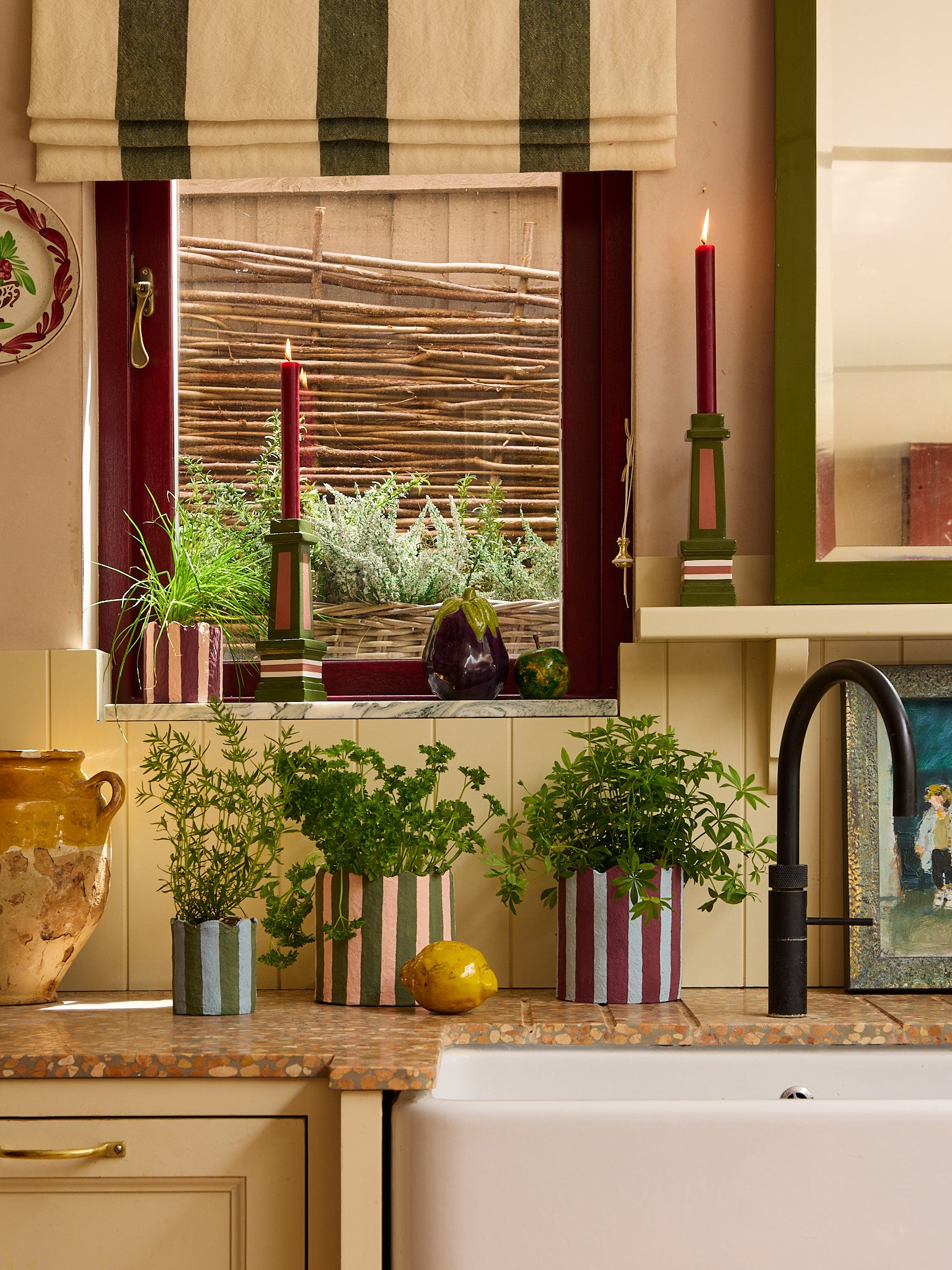 A kitchen counter with a sink and window, decorated with lit candles and must have planters